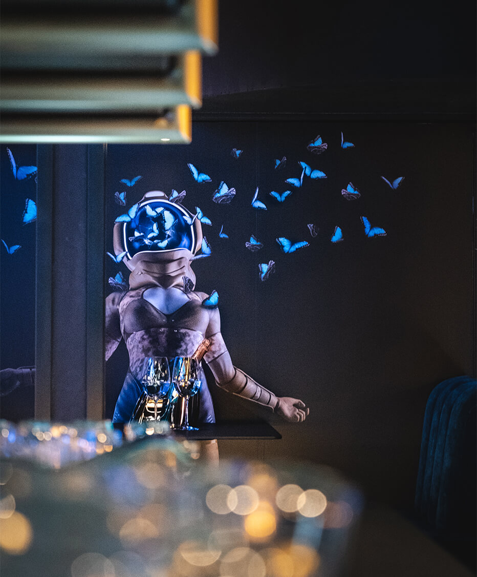 Wandgestaltung und Dekoration von der id Werkstatt aus Vorchdorf - Astronautin mit Schmetterlinge an Wand tapeziert, eye catcher im Eingangsbereich vom Club Luna in Innsbruck. Unschraf im Vordergrund Cocktailgläser und Flaschenregal aus Messing