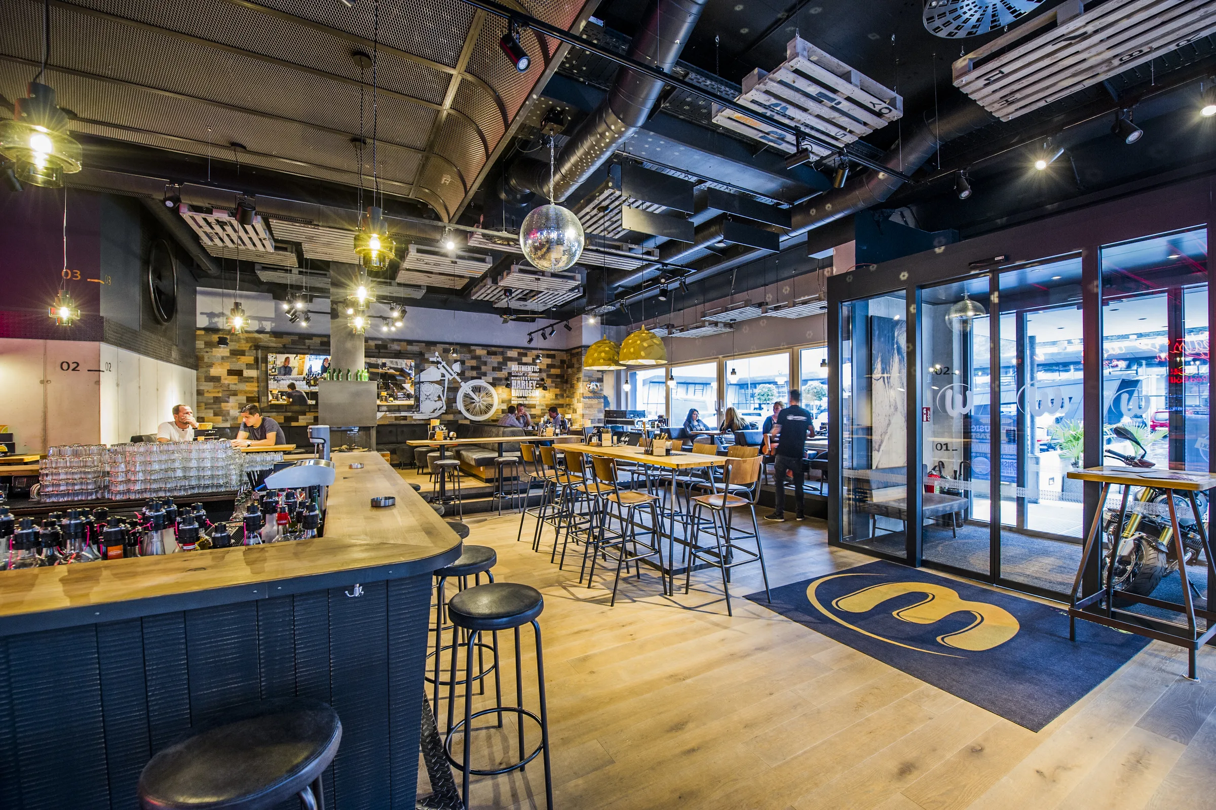 Bar und Gastraum der Werkstatt Kufstein mit dunklem Bartresen, schwarzen Barhockern, langen Stehtischen, Discokugel, Harley-Davidson-Wandbild und Logo-Eingangsmatte beim Glaseingang.
