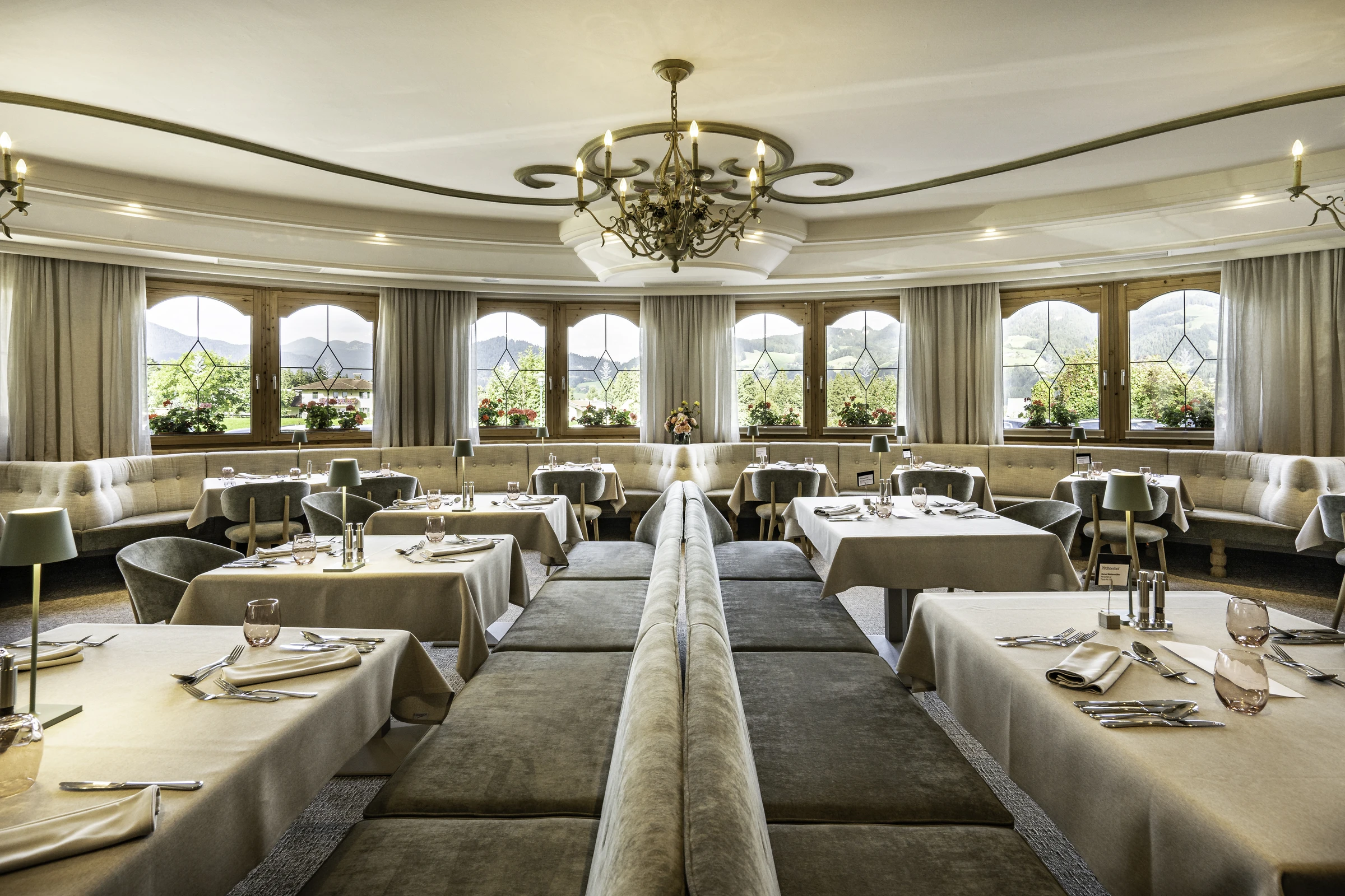 Restaurant des Pirchnerhof in Reith im Alpbachtal mit rundem Gastraum, eleganter Polstersitzbank, gedeckten Tischen mit Tischlampen, bronzenem Kronleuchter, Bleiglasfenstern mit Blumendekor und Panoramablick auf die Tiroler Berglandschaft.