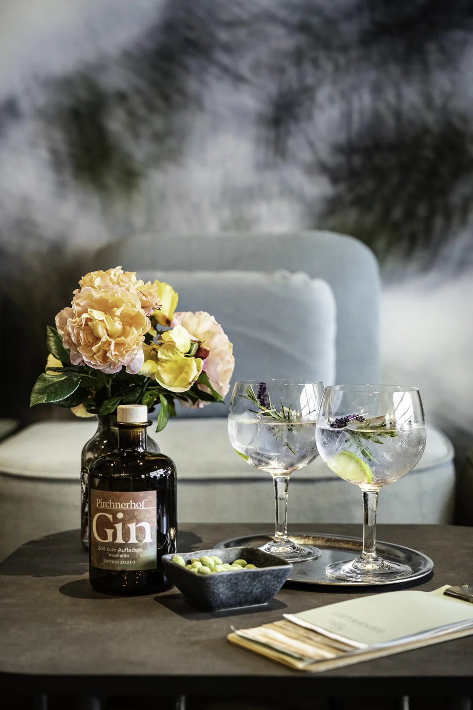 Detailaufnahme einer Pirchnerhof-Gin-Flasche mit Eigenlabel neben zwei Gin-Tonic-Gläsern mit Kräutern und Limette sowie einem Blumengesteck auf dem Beistelltisch der Lounge im Pirchnerhof Reith.