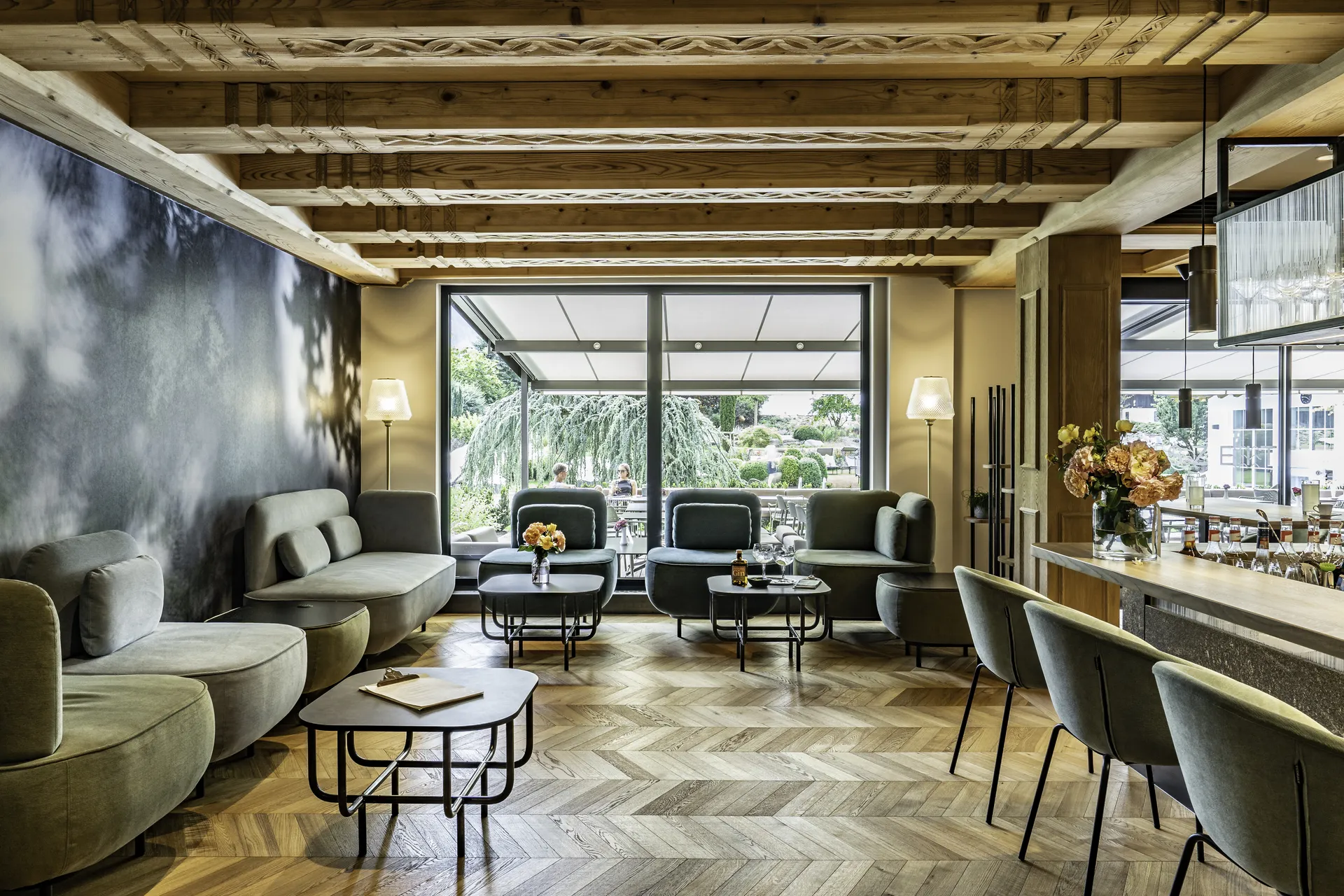 Lounge und Bar des Pirchnerhof Reith im Alpbachtal mit grünen Samtsesseln, geschwungenem Sofa, geschnitzter Altholzbalkendecke, Fischgrätparkett, Stehlampen, Panoramafenstern zur Terrasse und Blumengesteck an der Bar.