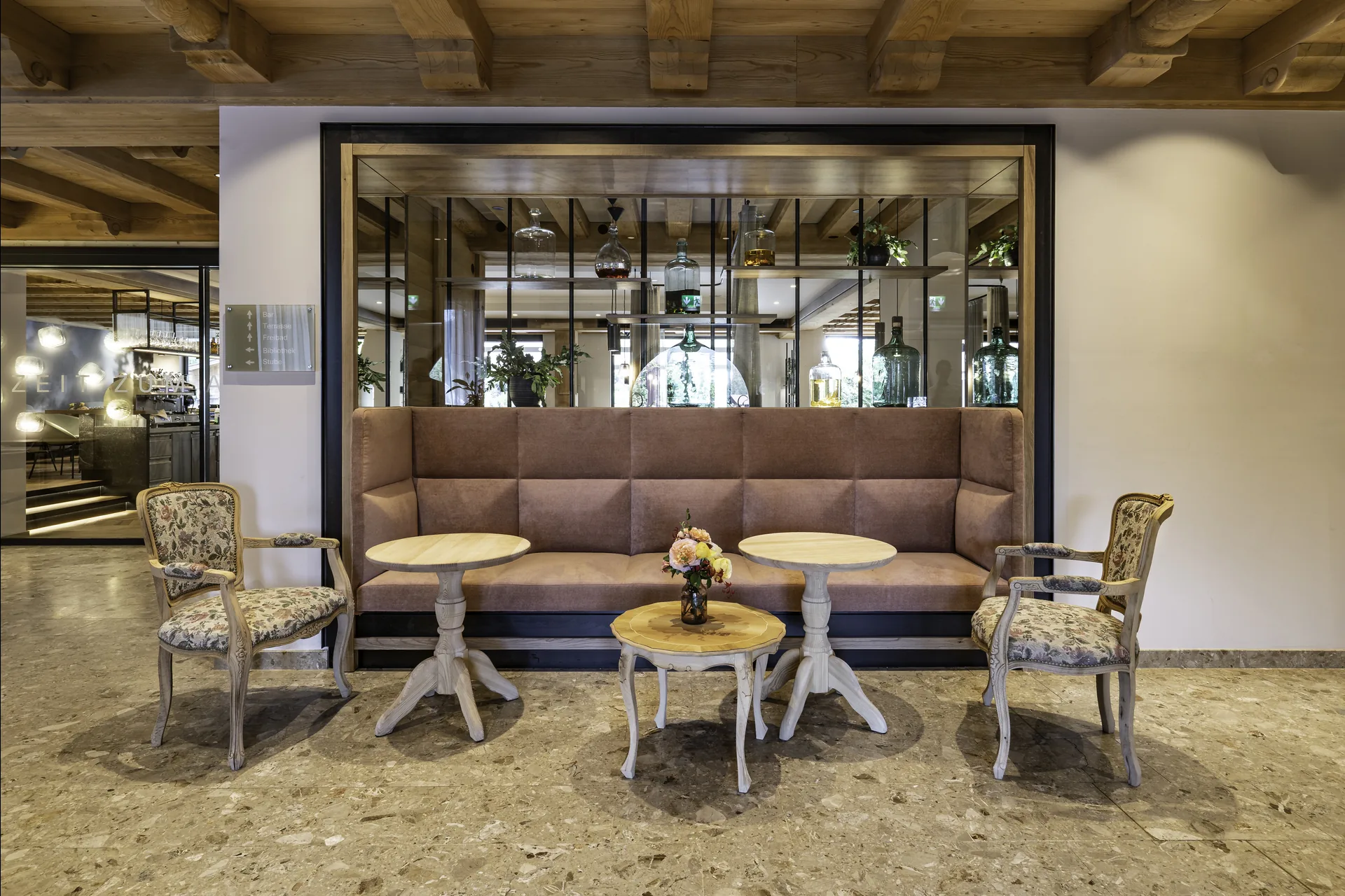 Lobby im Pirchnerhof Reith mit brauner Sitzbank, geblümten Sesseln, Rundtischen, Glasregal mit Spirituosen und Altholzdecke.