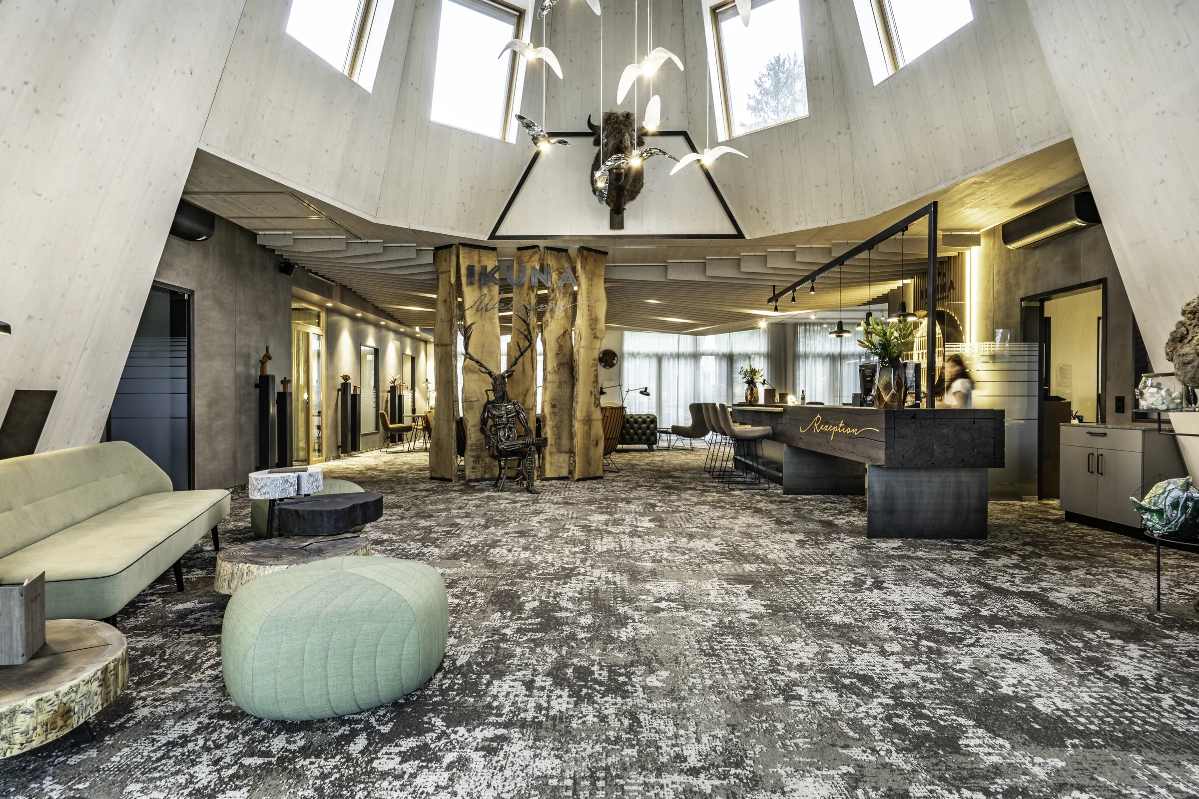 Lobby des IKUNA Tipi Hotel Natternbach mit Tipi-förmiger Holzkonstruktion, markanten Baumstämmen mit IKUNA-Schriftzug, einer Skulptur, mintgrünem Sofa und Pouf, dunklem Rezeptionstresen und großformatigem Vintageteppich.