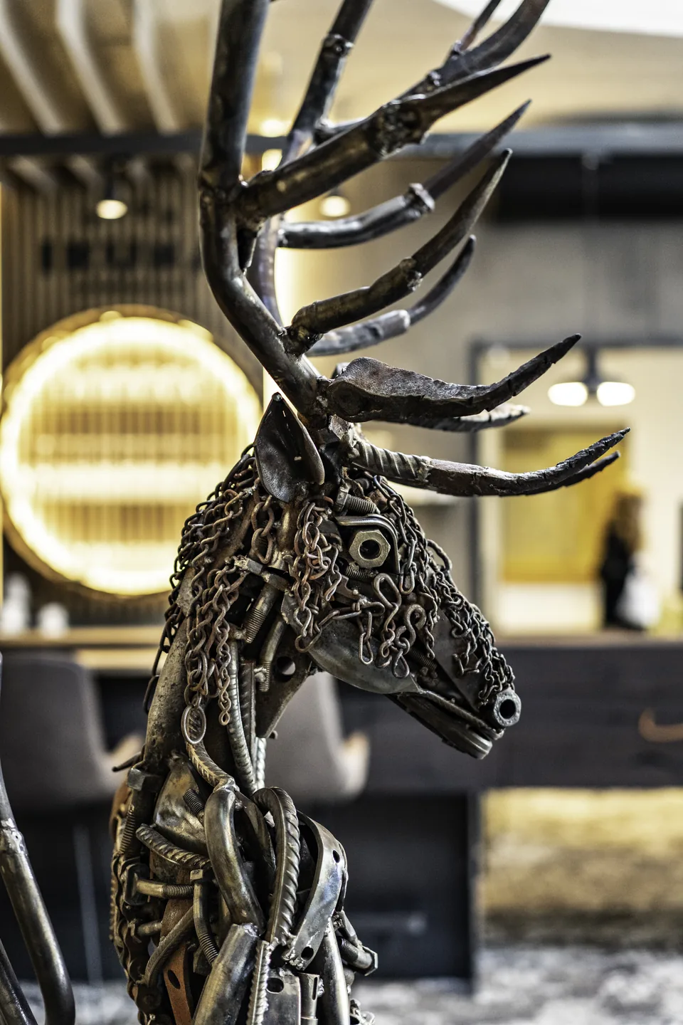 Detailaufnahme einer lebensgroßen Hirschskulptur aus Metallschrauben, Ketten und recycelten Metallteilen in der Lobby des IKUNA Tipi Hotel Natternbach, im Hintergrund der beleuchtete Rezeptionsbereich.