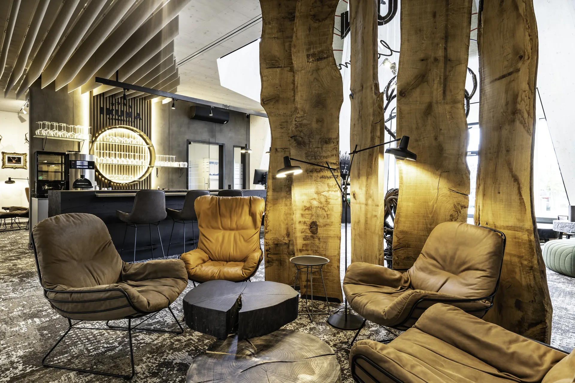 Loungebereich im IKUNA Tipi Hotel Natternbach mit cognacfarbenen Ledersesseln, einem Couchtisch aus Baumscheibe, beleuchteten Baumstämmen als Raumteiler, Vintageteppich und dem goldenen Barbereich mit rundem Glasregal im Hintergrund.