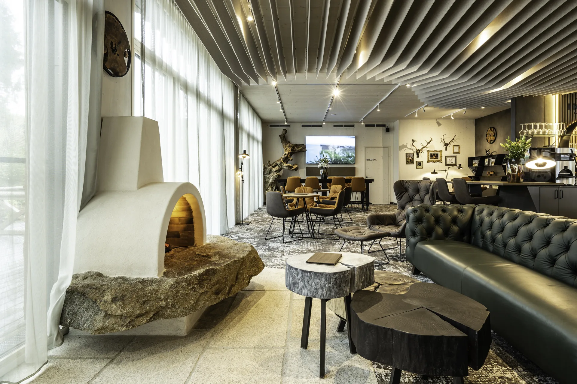 Loungebereich im IKUNA Tipi Hotel Natternbach mit einem freistehenden Lehmkamin auf Natursteinsockel, dunkelgrünem Chesterfield-Sofa, Baumscheiben-Couchtischen, Vintageteppich und dem Restaurantbereich mit Wildtrophäen im Hintergrund.