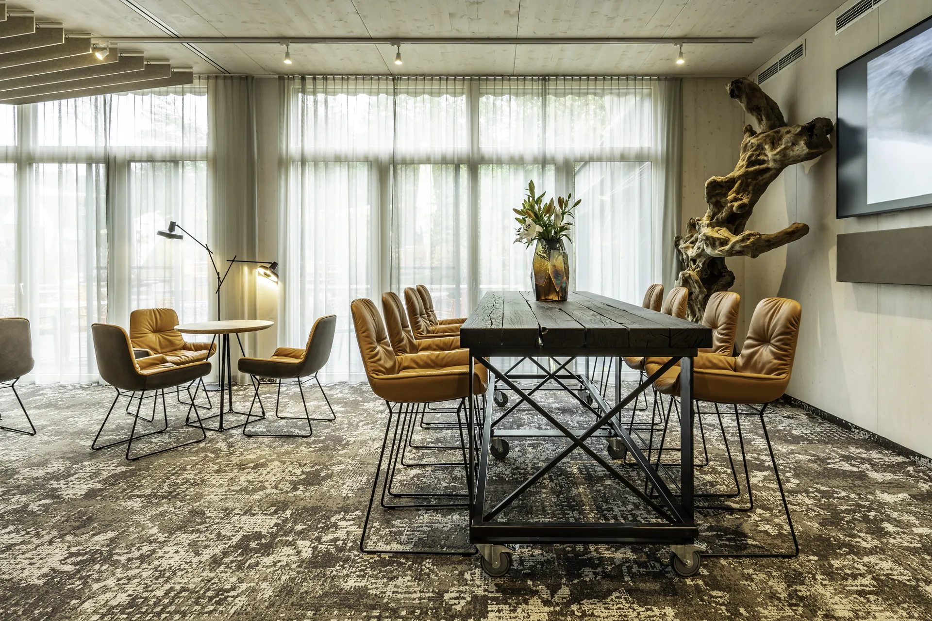 Seminar- und Meetingbereich im IKUNA Tipi Hotel Natternbach mit einem langen Holztisch auf Industrierollen, cognacfarbenen Ledersesseln, Vintageteppich, einer Treibholzskulptur an der Wand und bodenlangen Vorhängen.