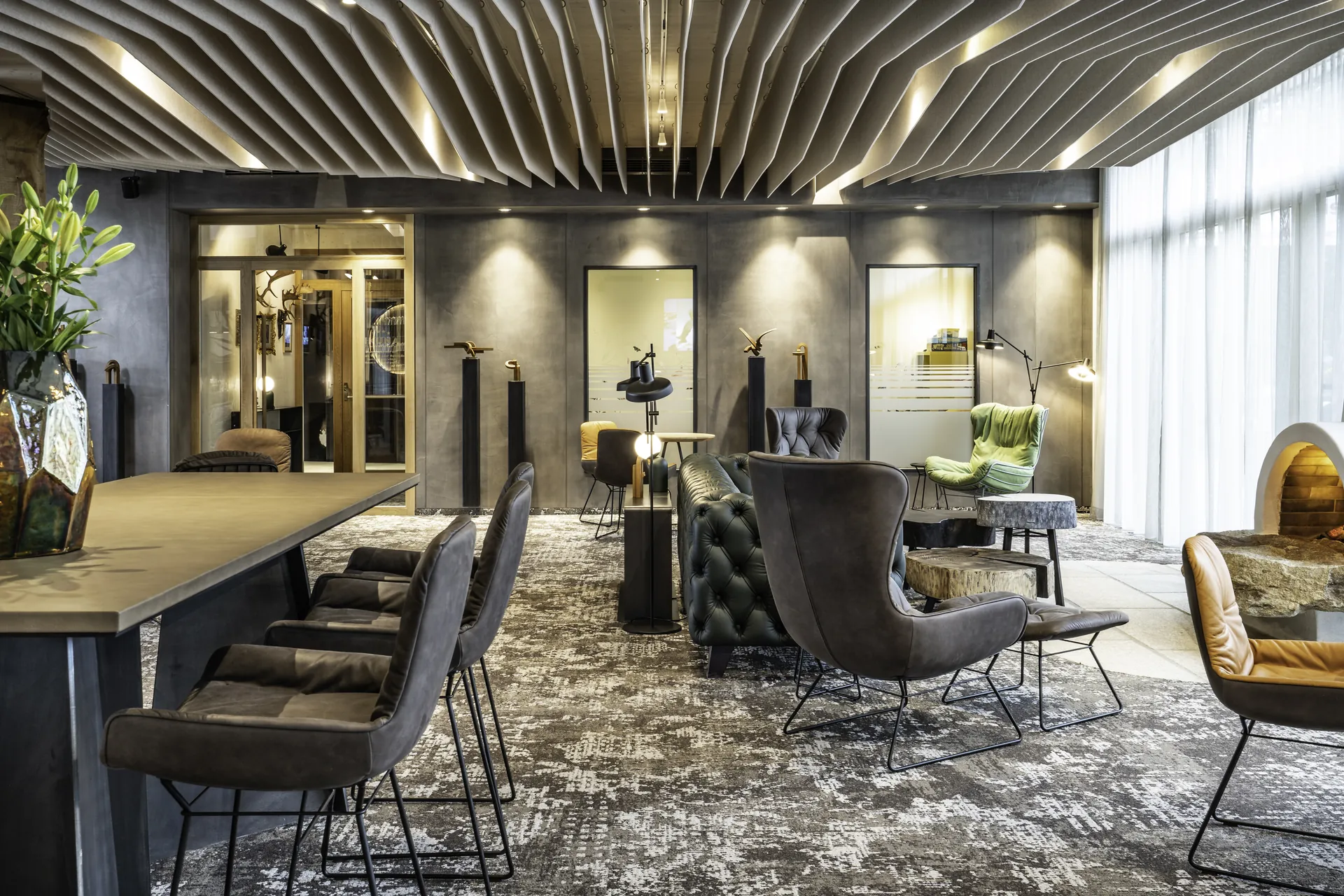 Loungebereich im IKUNA Tipi Hotel Natternbach mit verschiedenen Sesseln und einem dunkelgrünen Chesterfield-Sofa, Vintageteppich, markanter Wellendecke, bronzefarbenen Tierskulpturen auf Sockeln und warmem Licht.