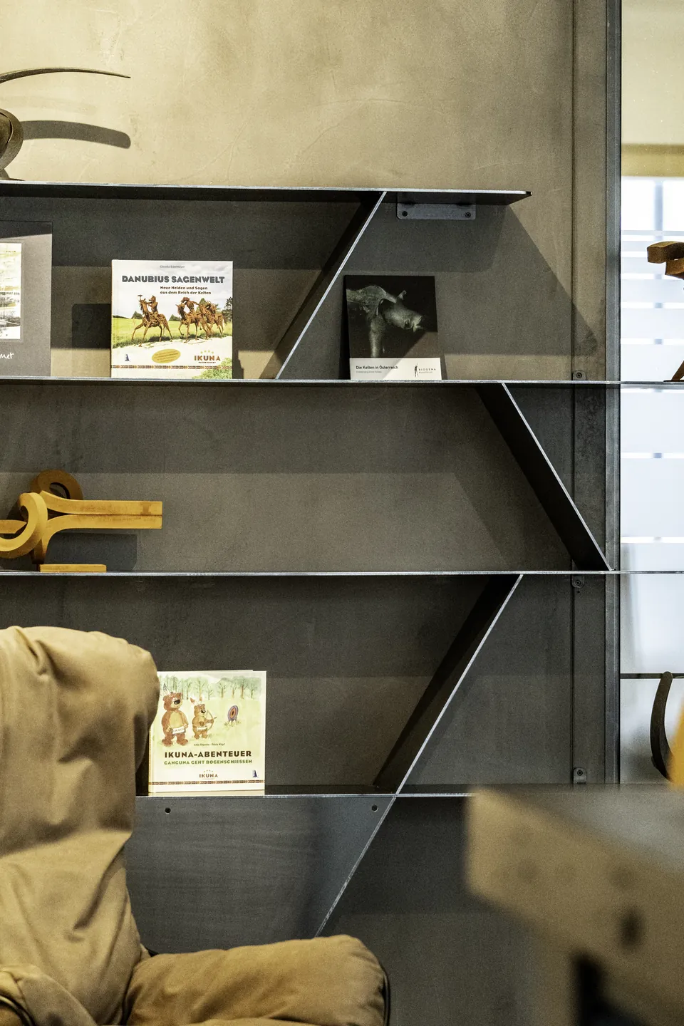 Detailaufnahme eines Metallregals in der Lobby des IKUNA Tipi Hotel Natternbach mit IKUNA-Büchern, einem Kinderbuch, einer Holzskulptur und einem Foto vor einer dunklen Betonwand.