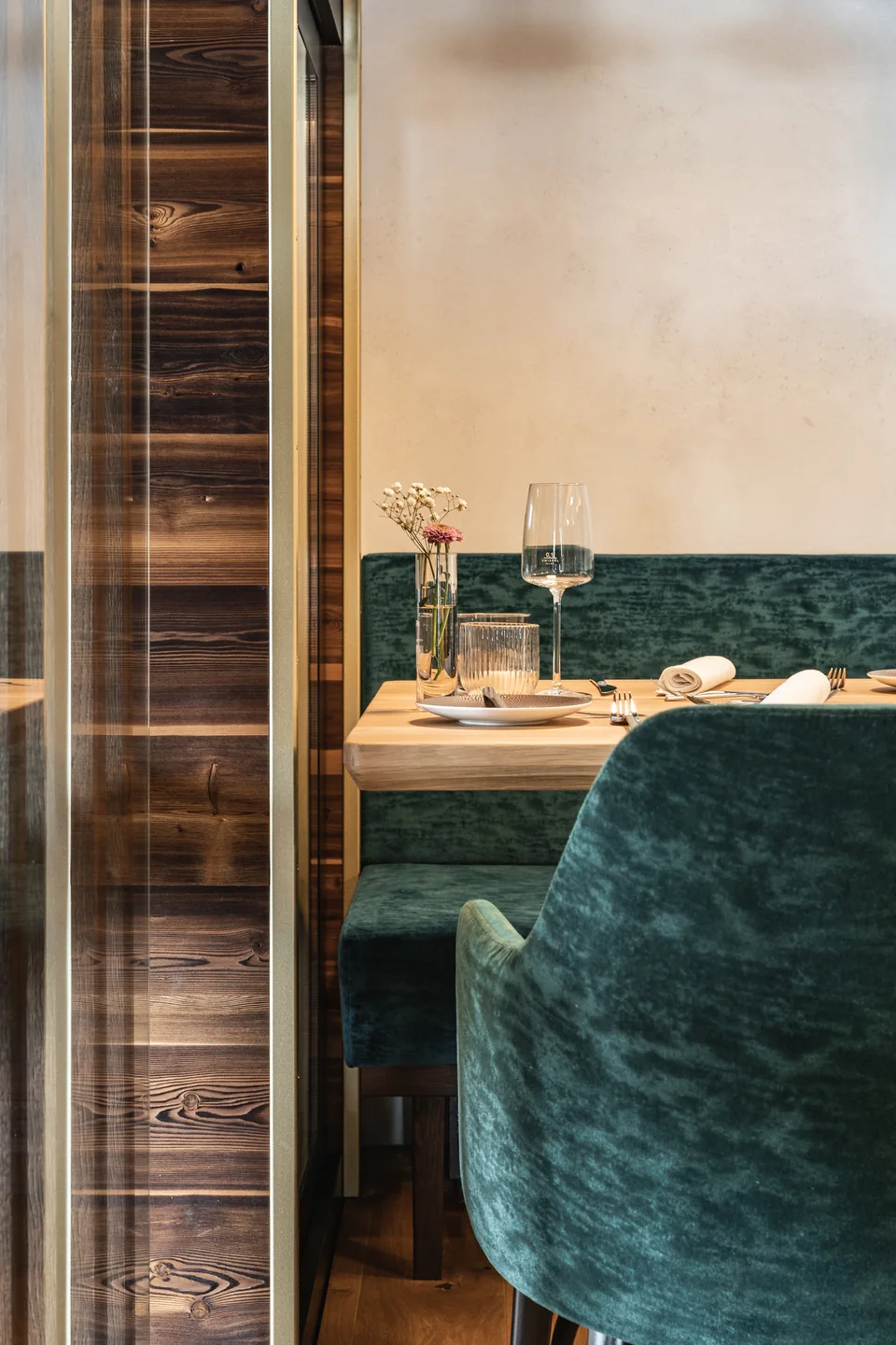 Gedeckter Tisch im Restaurant Blauer Bock Passau mit türkisfarbenen Samtsesseln, Weinglas, Blumenvase und dunkler Holzvertäfelung hinter einer Glasscheibe.