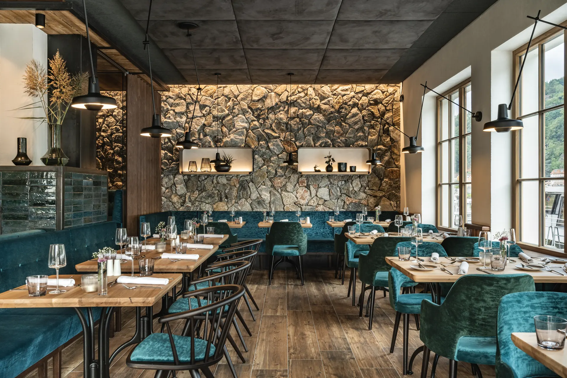 Vollständig gedeckter Gastraum des Restaurants Blauer Bock Passau mit türkisfarbenen Samtstühlen, gedeckten Holztischen, beleuchteter Natursteinwand und großen Panoramafenstern.
