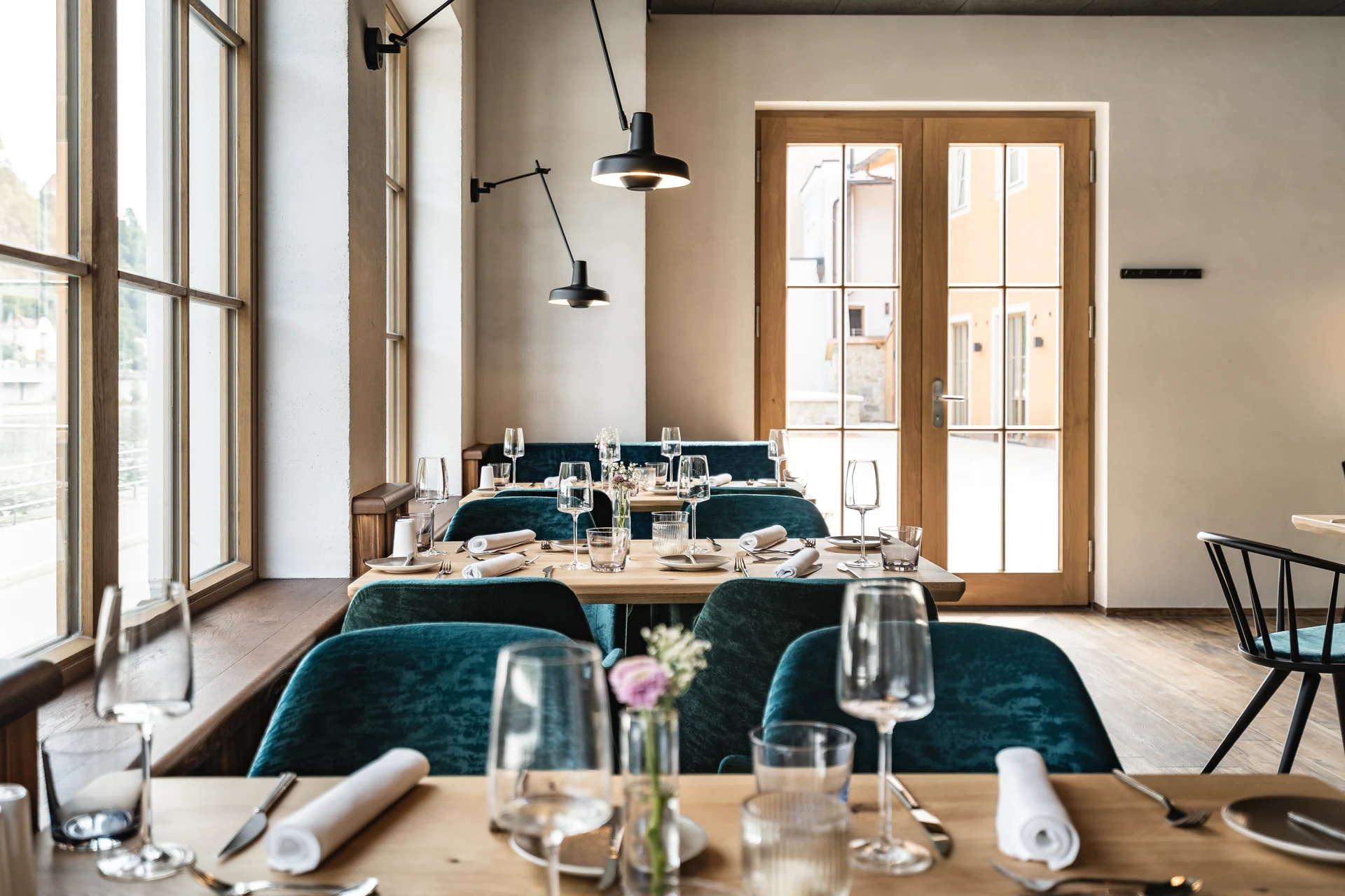 Heller Gastraum des Restaurants Blauer Bock Passau mit gedeckten Holztischen, türkisfarbenen Samtstühlen, schwarzen Wandleuchten und Blick durch die Terrassentür auf die Altstadt.