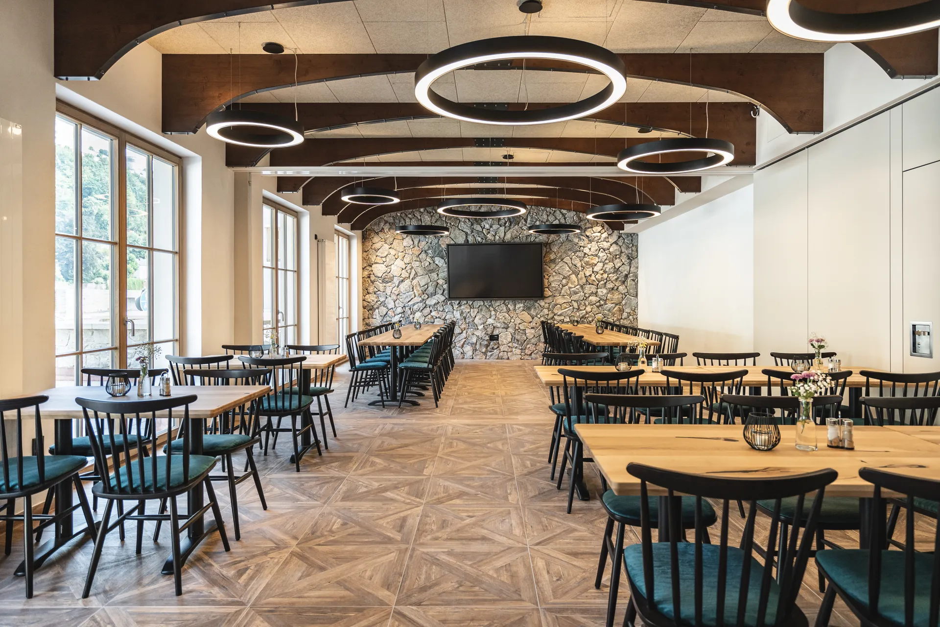 Großer Festsaal des Restaurants Blauer Bock Passau mit schwarzen Holzstühlen, Holztischen, markanten Ring-Hängeleuchten, Altholzdeckenbalken und Natursteinwand mit Bildschirm.