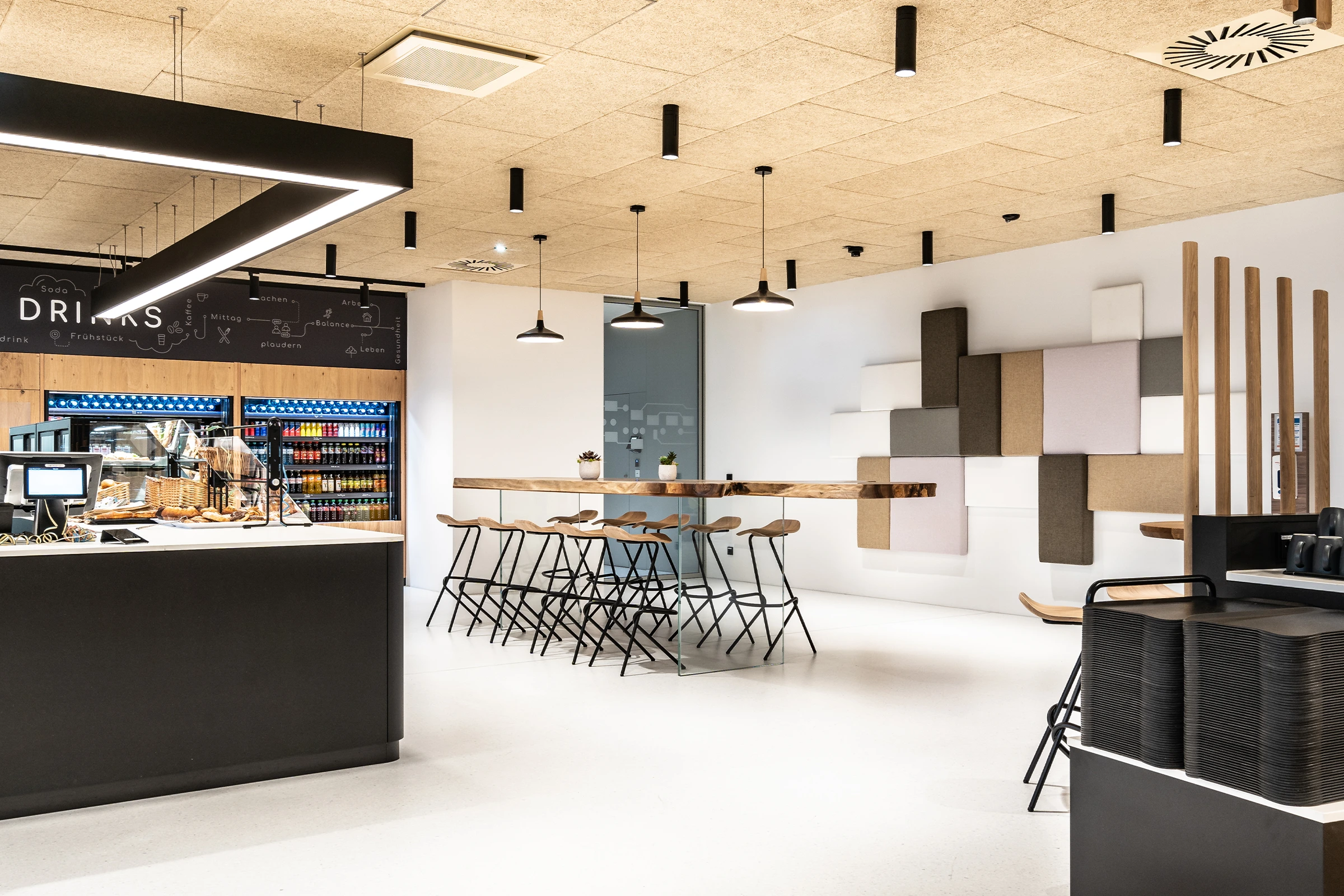 Moderner Ausgabebereich eines Mitarbeiterrestaurants mit schwarzer Theke, beleuchtetem Getränkekühlregal und einem Hochtisch mit Barhockern vor einer geometrischen Akustikwand.