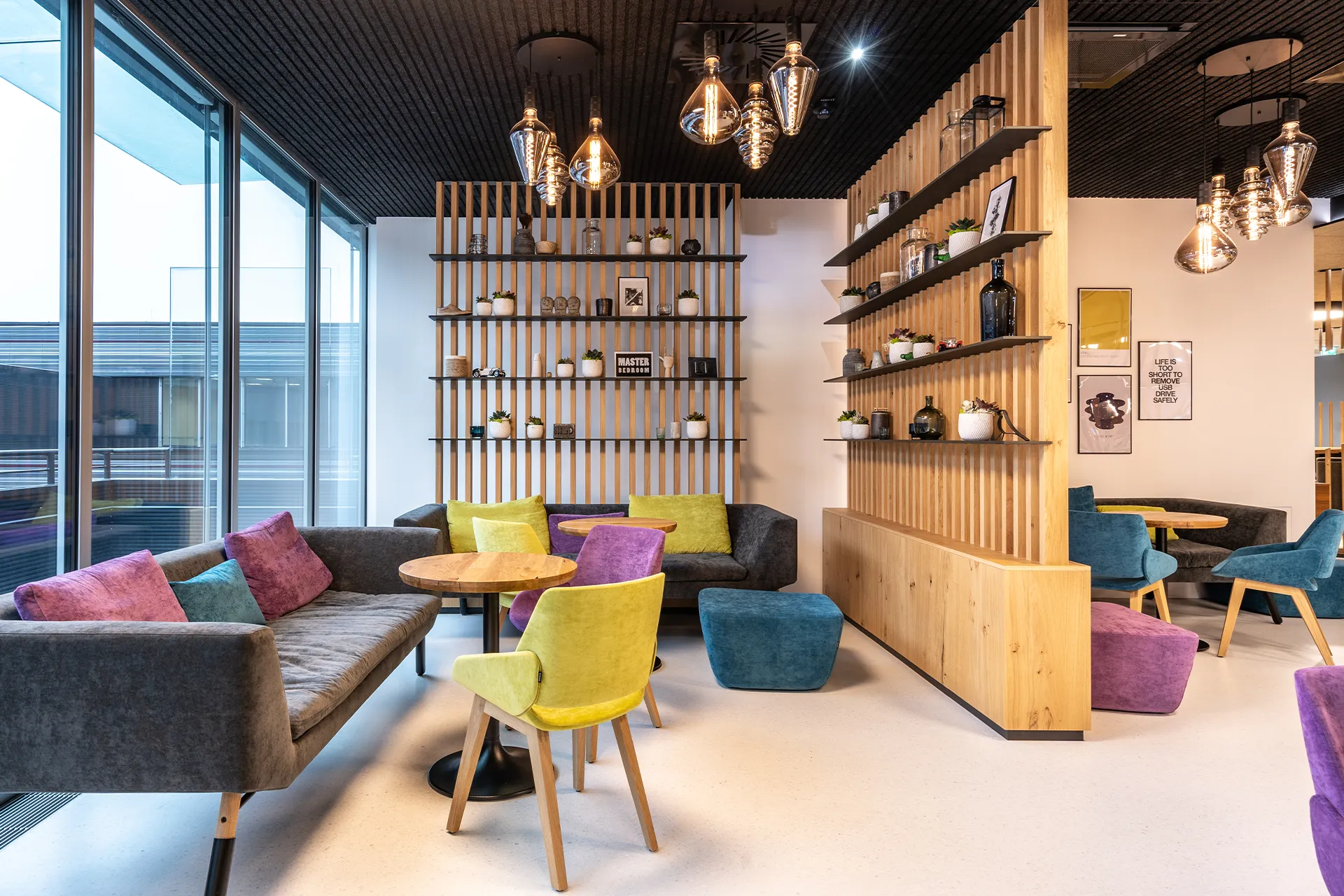 Gemütliche Lounge-Zone mit bunten Samtsesseln und -sofas, Holzlatten-Regalwand mit dekorativen Objekten und verspiegelten Pendelleuchten.