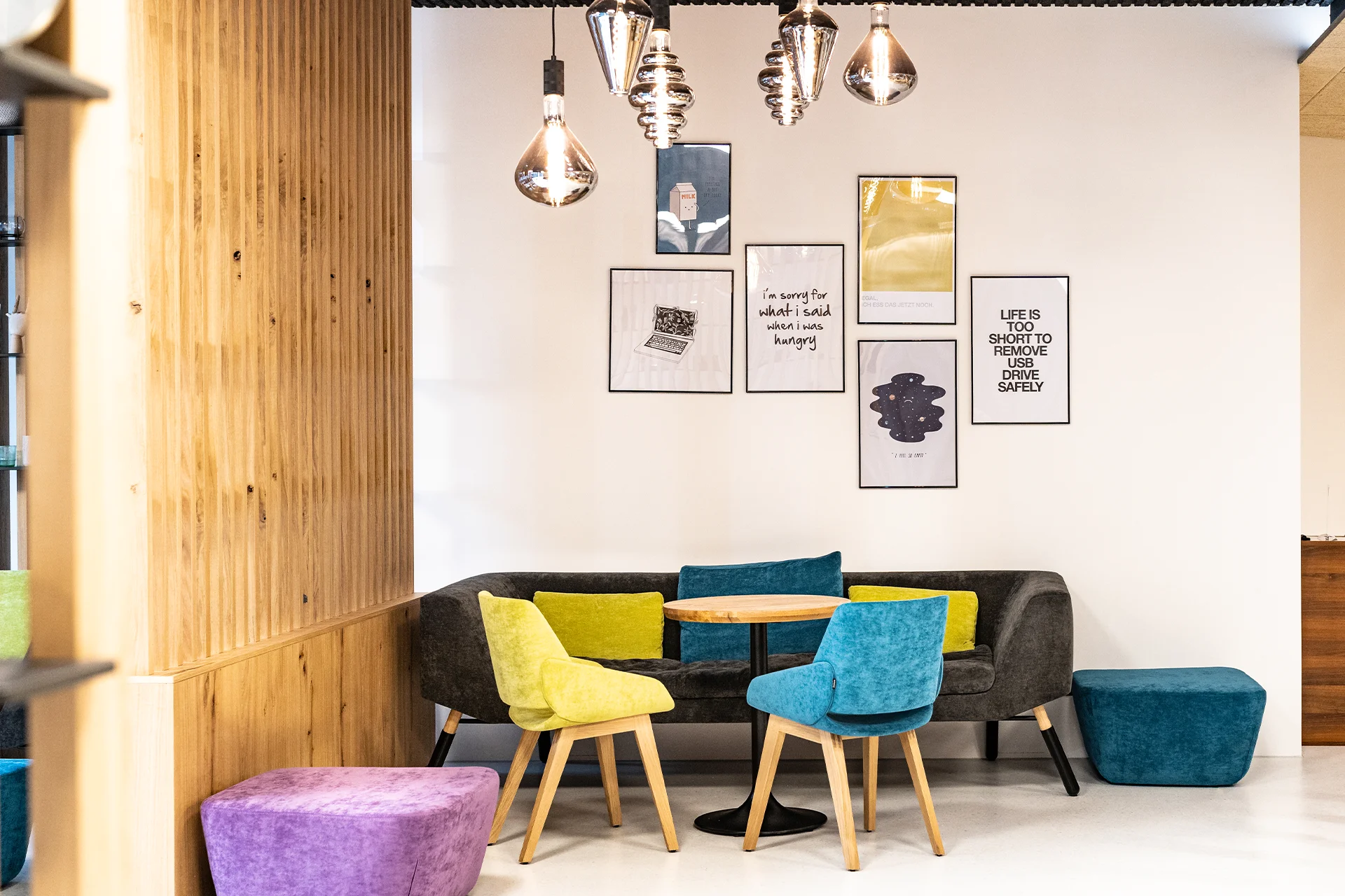 Entspannter Sitzbereich mit grauer Couch, bunten Stühlen und Hockern sowie einer Galeriewand mit humorvollen Typografie-Postern und Illustrationen.