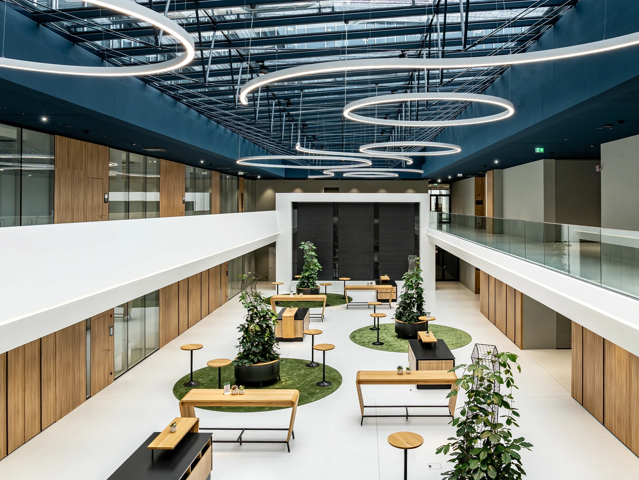 Atrium des BR Forum Eggelsberg aus der Vogelperspektive mit Holzstehtischen, Pflanzensäulen auf grünen Teppichinseln, umlaufender Galerie mit Glasbrüstung und ringförmigen Hängleuchten an blauer Decke