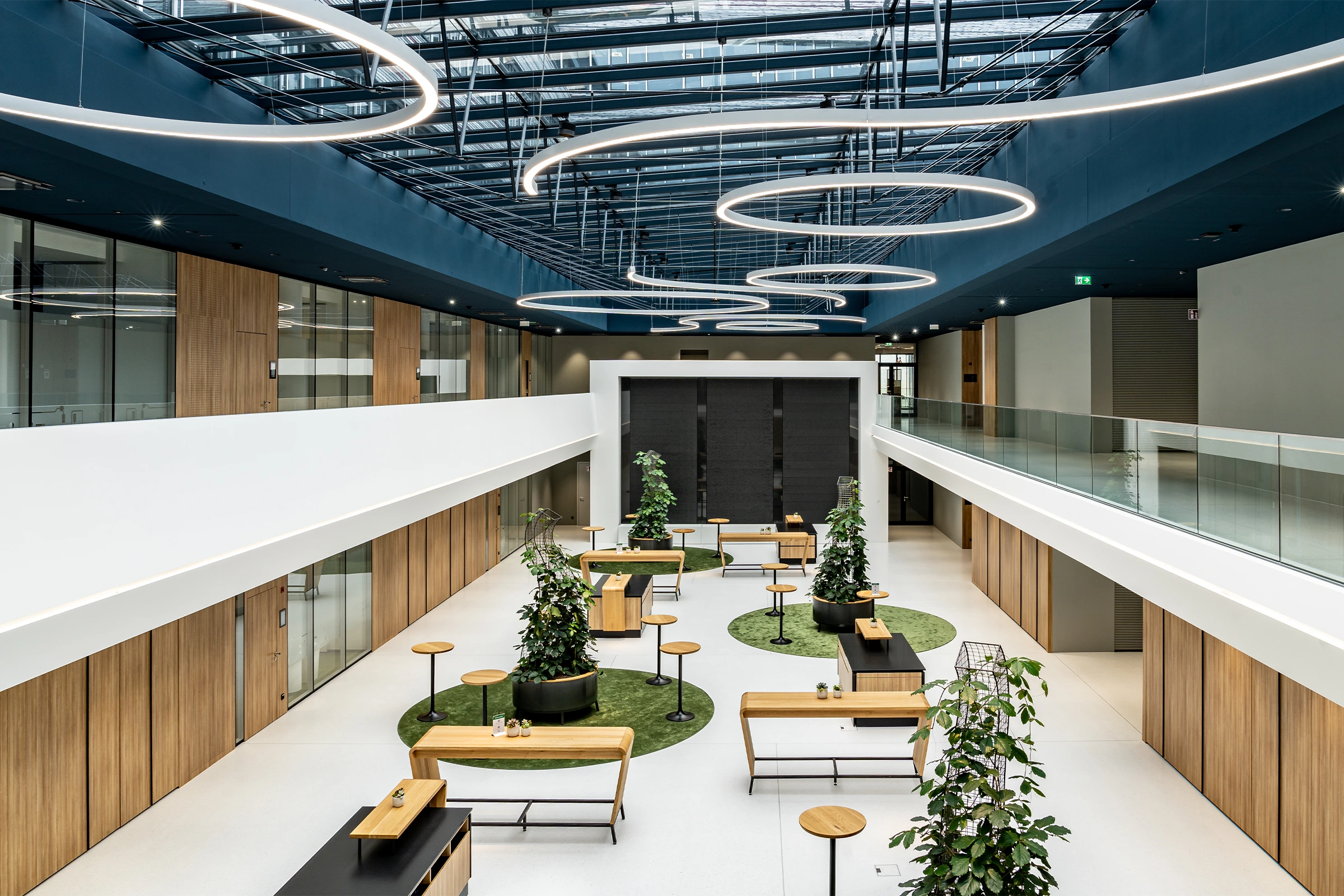 Atrium des BR Forum Eggelsberg aus der Vogelperspektive mit Holzstehtischen, Pflanzensäulen auf grünen Teppichinseln, umlaufender Galerie mit Glasbrüstung und ringförmigen Hängleuchten an blauer Decke