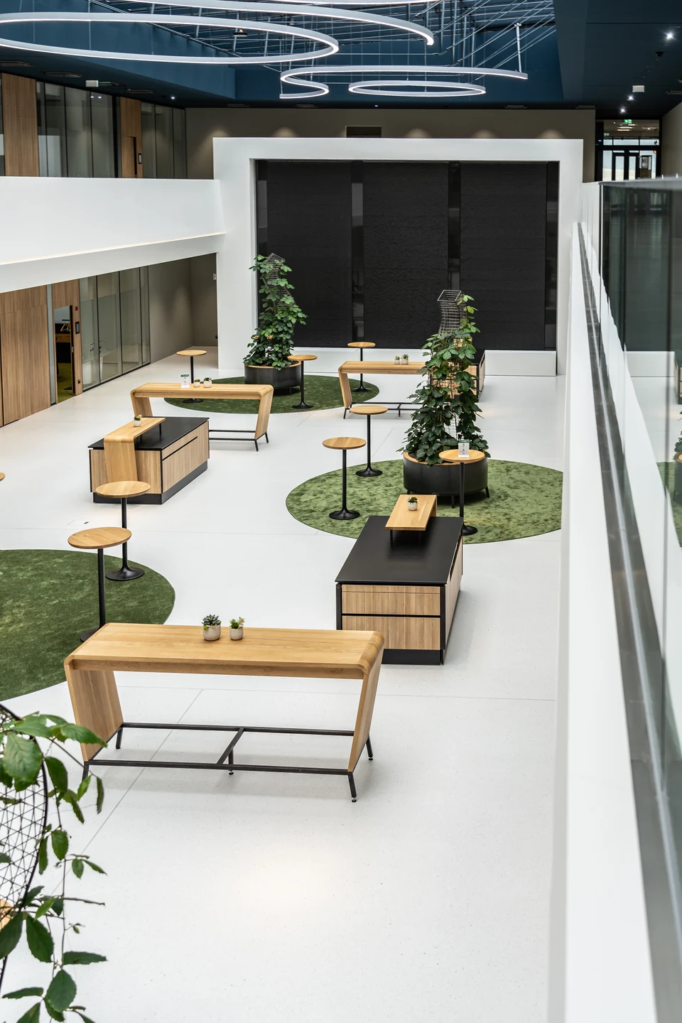 Atrium des BR Forum Eggelsberg aus der Vogelperspektive mit Holzstehtischen, Pflanzensäulen auf grünen Teppichinseln und Ringleuchten.