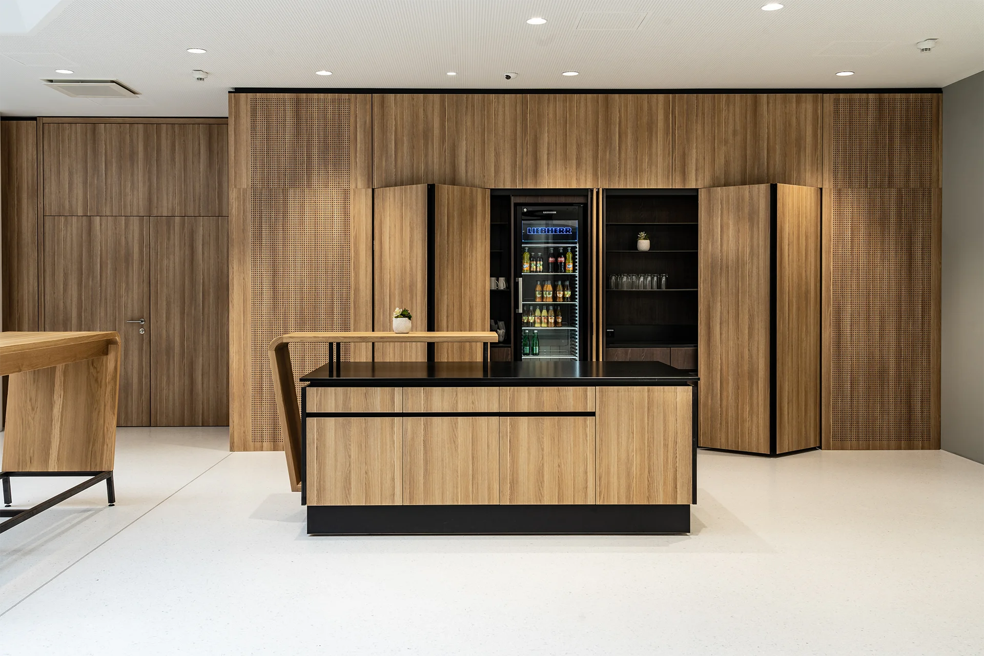 Elegante Empfangsbar aus Holz mit schwarzer Arbeitsplatte, integriertem Getränkekühlschrank und geöffneten Falttüren, die den Versorgungsbereich dahinter freilegen.