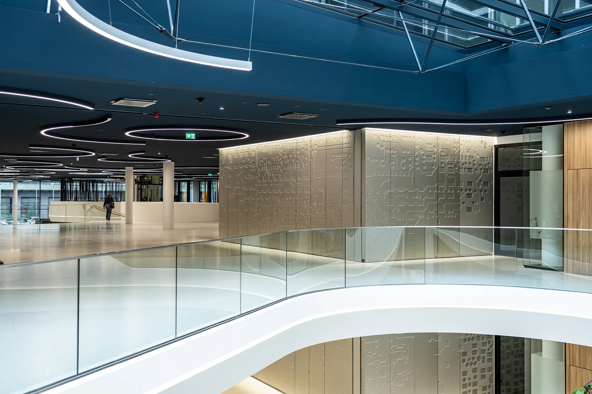 Blick von der Galerie auf einen mit Leiterplatten-Motiven gefrästen Wandkubus im Atrium, gerahmt von geschwungenen Glasbrüstungen und Ring-Leuchtinstallationen.