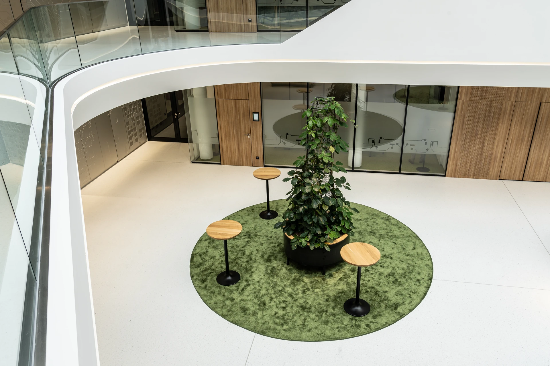 Atrium des BR Forum Eggelsberg mit zentraler Pflanzensäule, runden Stehtischen auf grünem Teppich und umlaufender Galerie.
