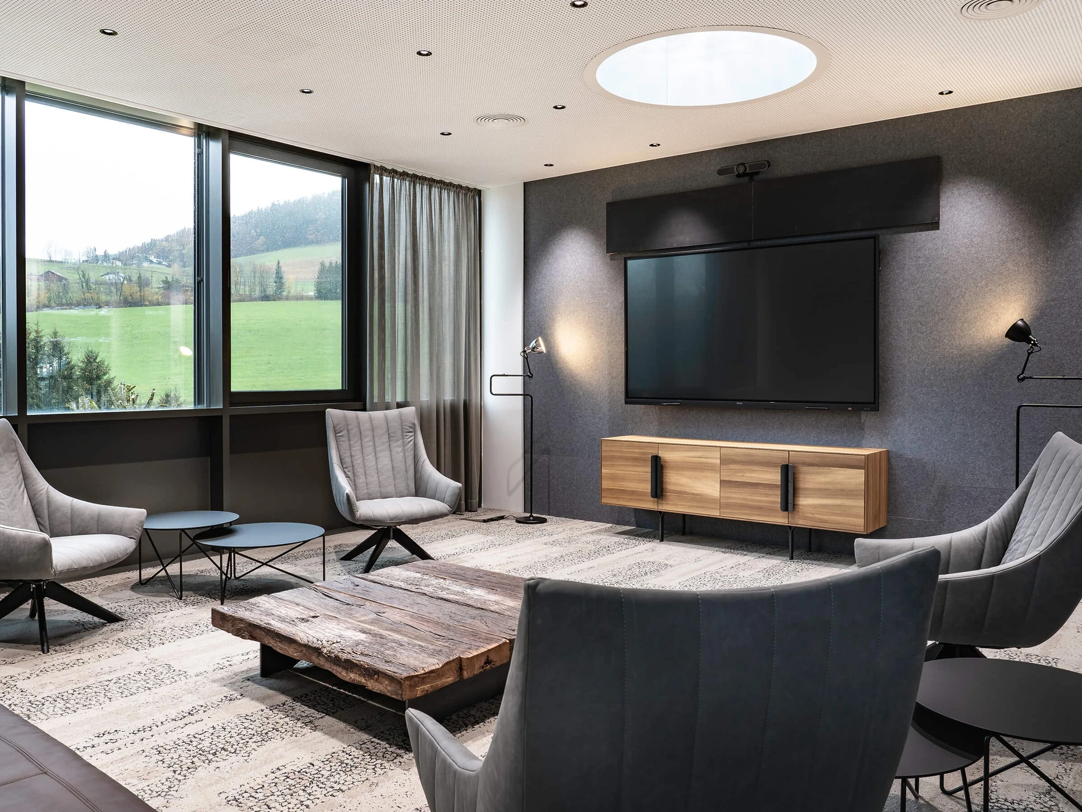 Loungebereich im Biogena Health World Büro Koppl mit grauen Sesseln, rustikalem Altholz-Couchtisch, Akustikwand mit Bildschirm und Panoramablick in die Salzburger Berglandschaft.
