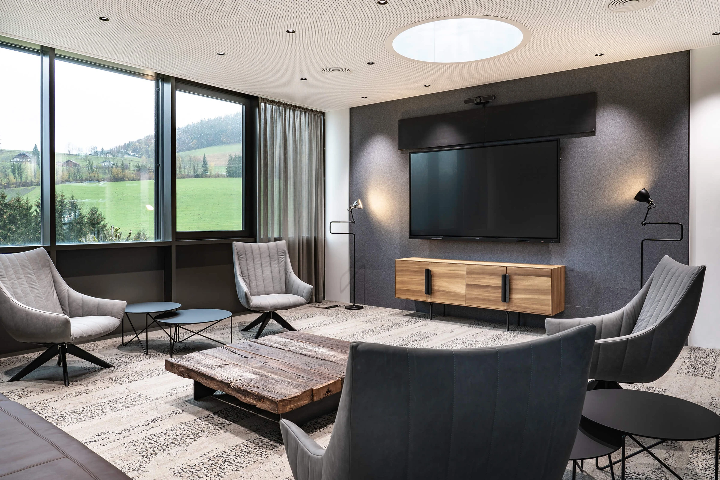 Loungebereich im Biogena Health World Büro Koppl mit grauen Sesseln, rustikalem Altholz-Couchtisch, Akustikwand mit Bildschirm und Panoramablick in die Salzburger Berglandschaft.