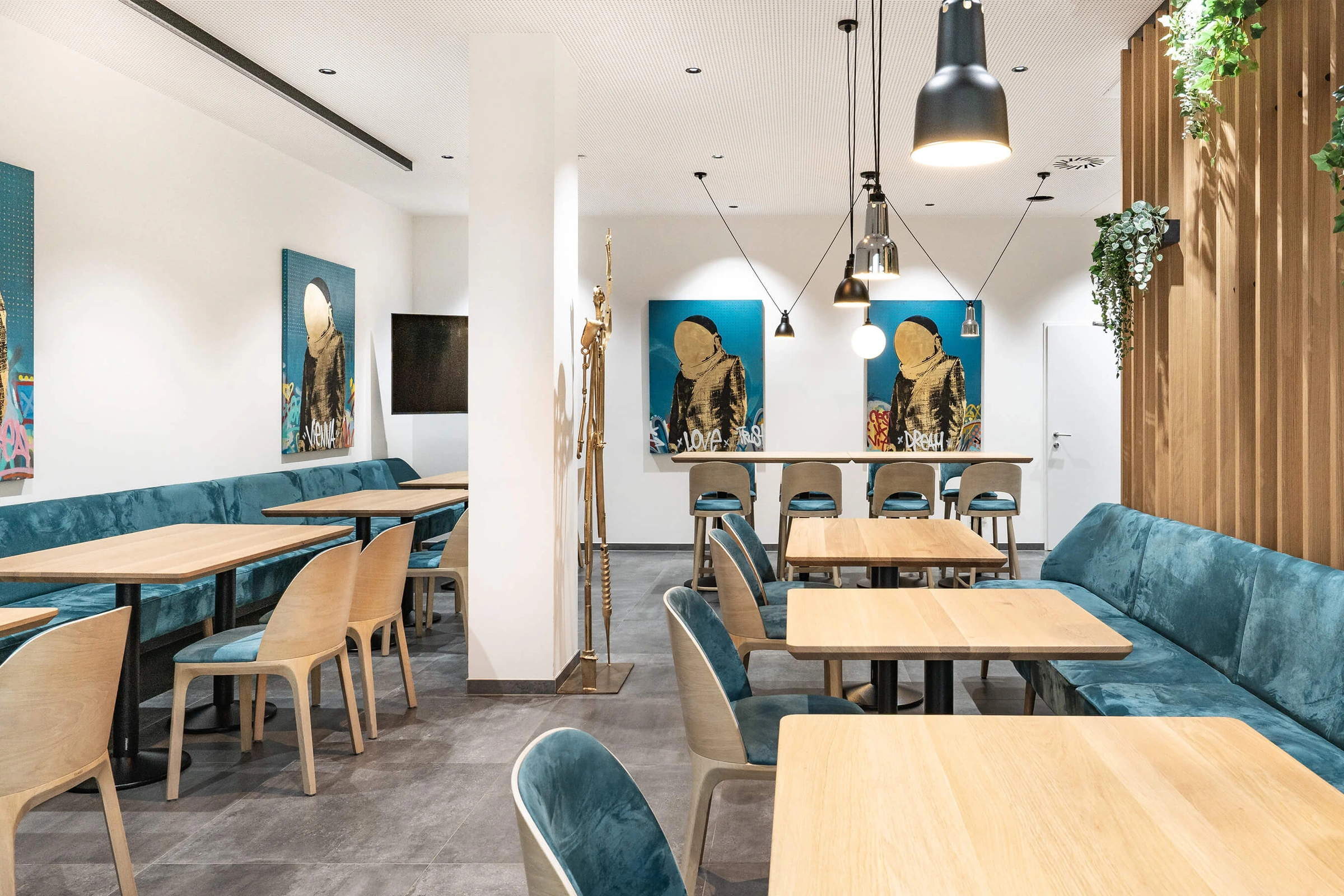 Bistro der Biogena Healthworld in Koppl mit blauer Samtpolsterbank, hellen Holztischen und -stühlen, auffälligen Street-Art-Kunstbildern in Blau-Gold, schwarzen Pendelleuchten, goldener Figurenskulptur und begrünten Holzlamellen.