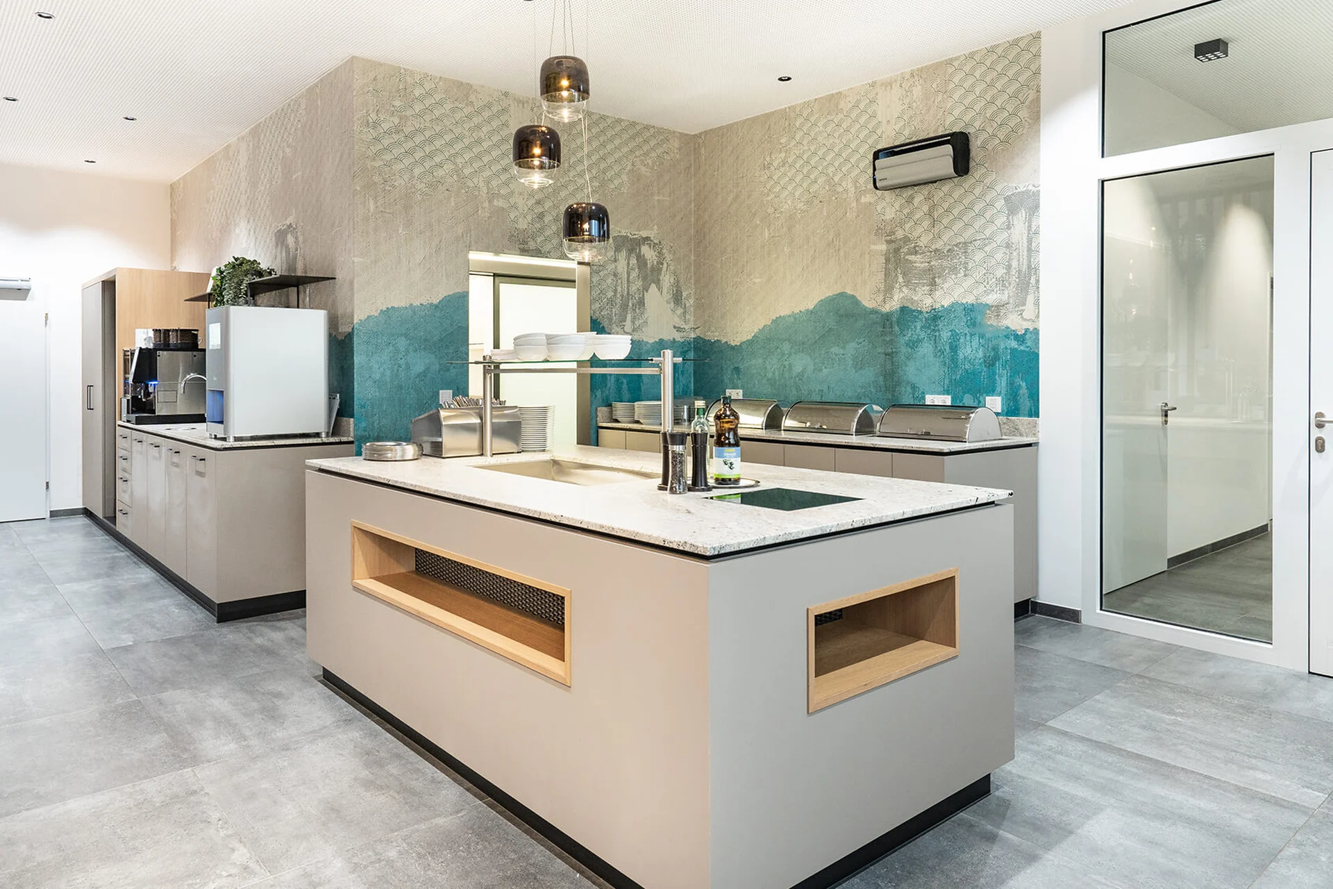 Bistroküche der Biogena Healthworld in Koppl mit zentraler Kochinsel mit Marmorarbeitsplatte, auffälliger türkis-grauer Designtapete, rauchigen Glashängeleuchten und Buffetstation im Hintergrund.