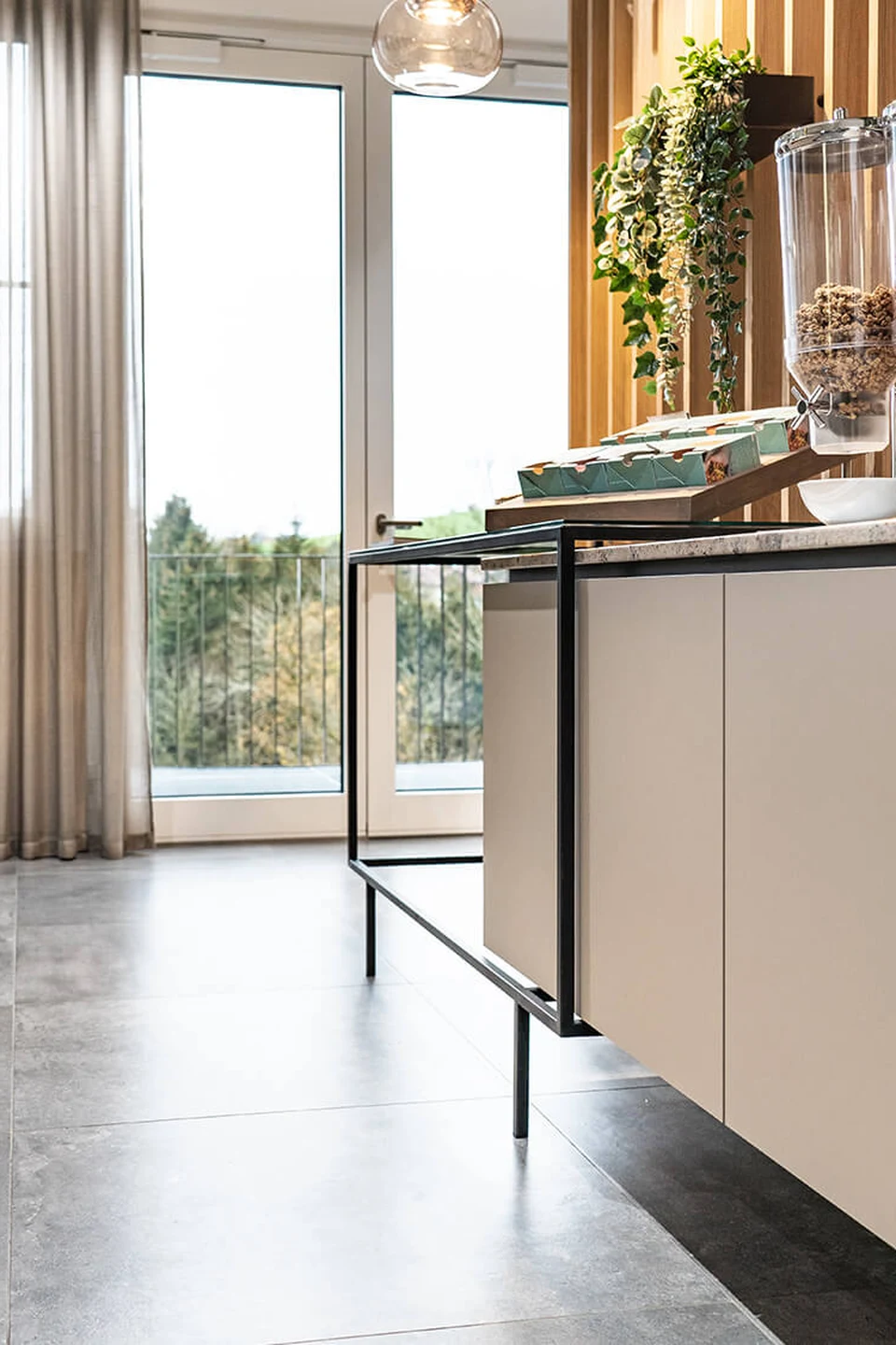 Detailaufnahme der Ausgabetheke im Bistro der Biogena Healthworld in Koppl mit Müslidispenser, Snackboxen, hängender Begrünung und Panoramafenster mit Blick auf Balkon und Baumlandschaft.