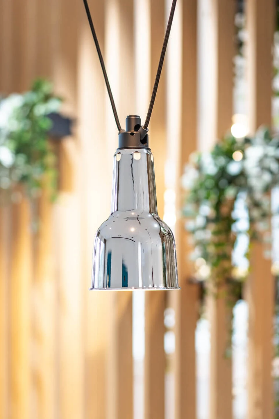 Detailaufnahme einer glänzenden Chrom-Pendelleuchte vor beleuchtetem Holzlamellenraster mit hängender Begrünung im Bistro der Biogena Healthworld in Koppl.