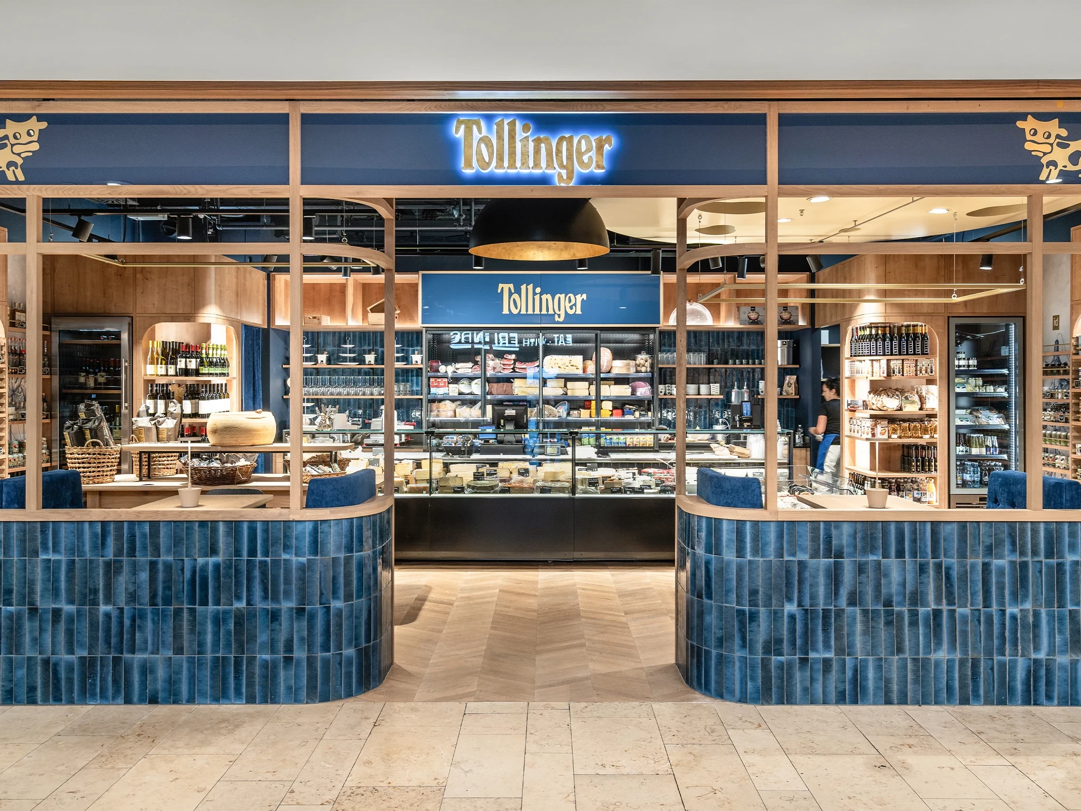 Ein modern gestaltetes Feinkostgeschäft der Marke Tollinger mit einer hölzernen Schaufensterfront, blauen Fliesenakzenten und einer gut sortierten Käse- und Weinpräsentation im Innenraum.