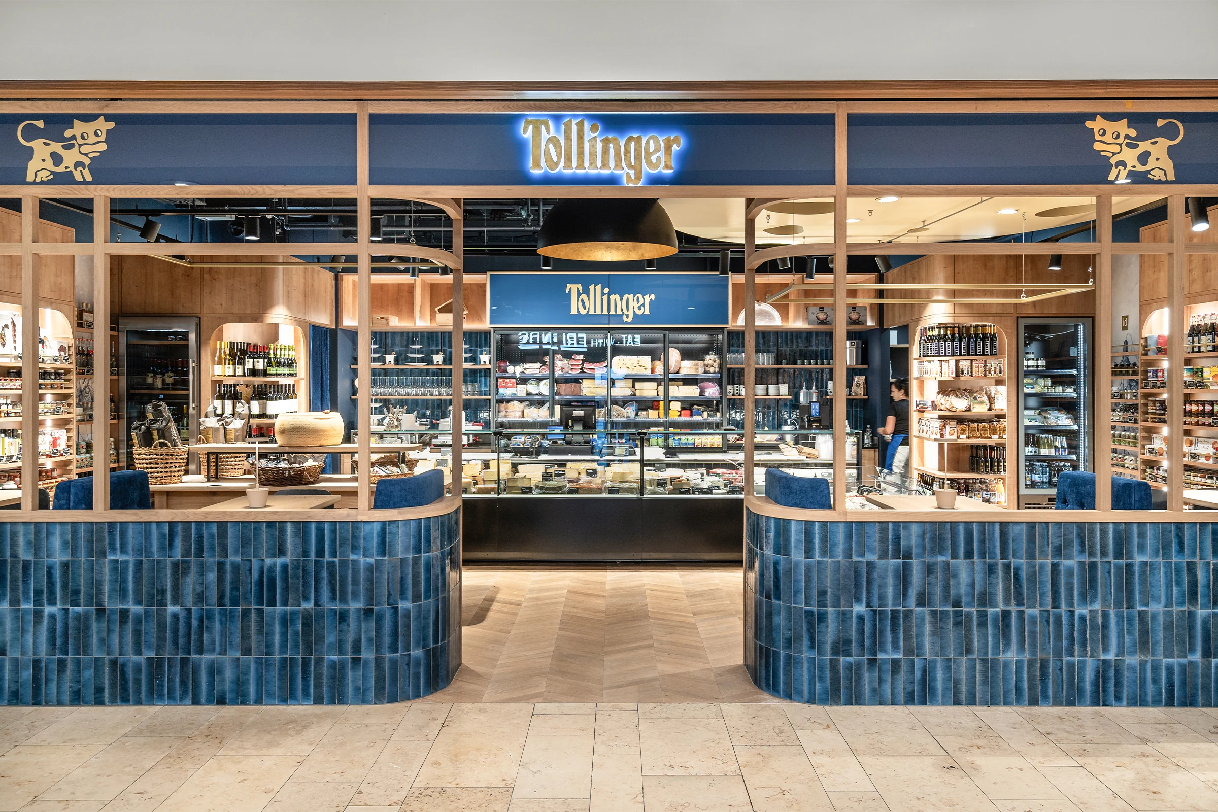 Ein modern gestaltetes Feinkostgeschäft der Marke Tollinger mit einer hölzernen Schaufensterfront, blauen Fliesenakzenten und einer gut sortierten Käse- und Weinpräsentation im Innenraum.