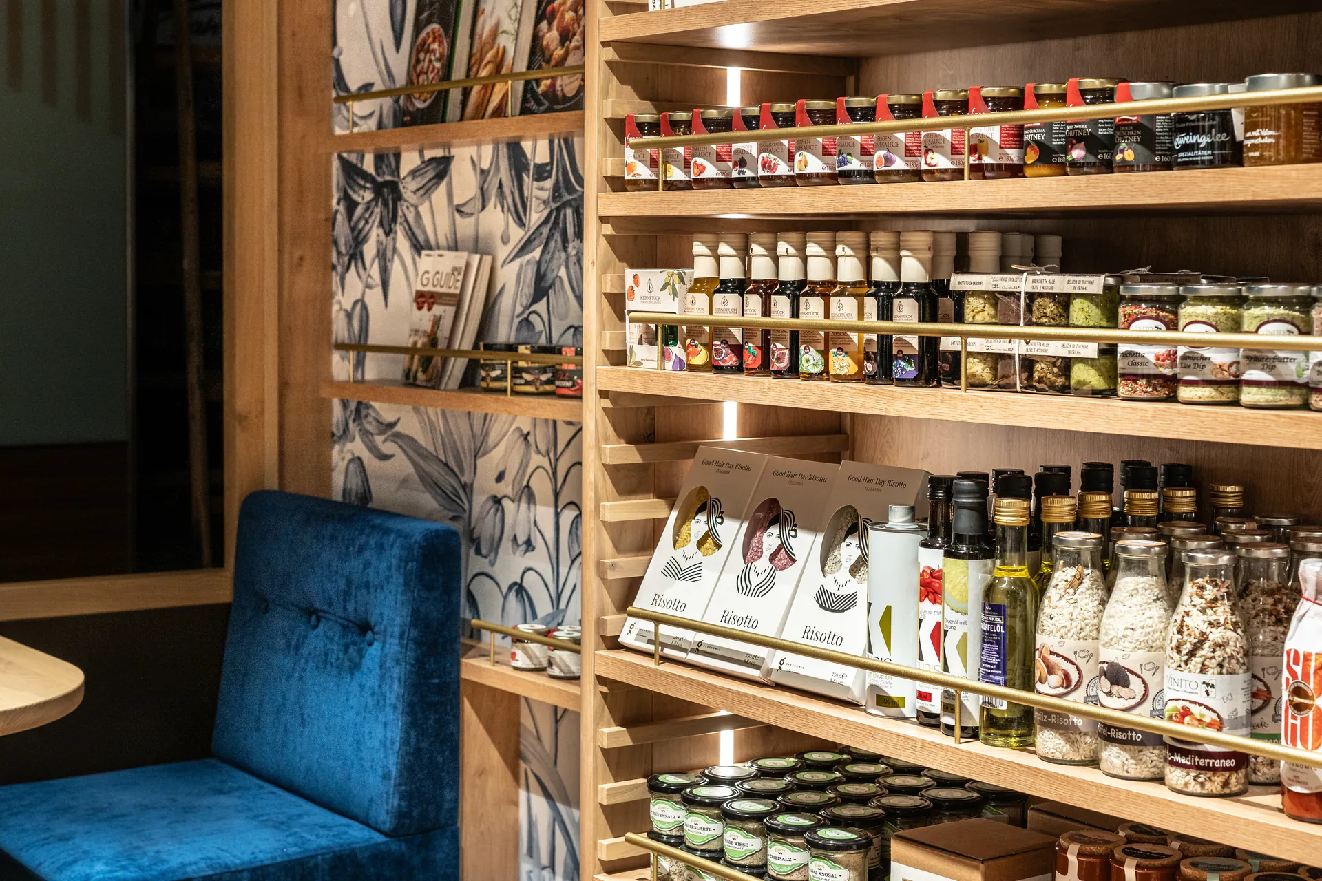 Holzregal mit Spezialitäten, Risotto, Öl und Konfitüren bei Tollinger Innsbruck, mit blauer Sitzbank und floraler Tapete im Hintergrund.