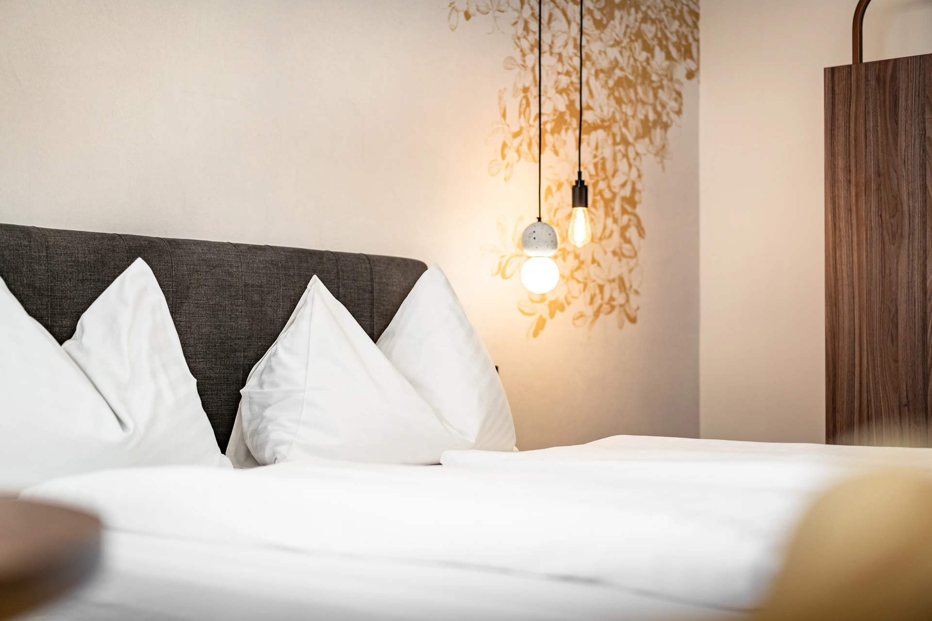 Detailaufnahme eines Hotelbetts im Hotel Lokomotive Linz mit weißen Kissen, dunkelgrauem Polsterkopfteil, zwei hängenden Edison-Pendelleuchten und goldenem Blättermotiv an der Wand.