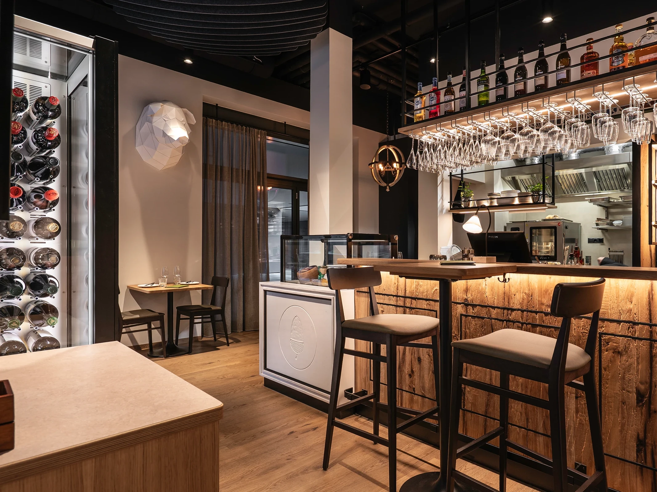 Bar des Paco Ribera mit beleuchtetem Weinkühlschrank, Barhockern am Naturholztresen, hängenden Weingläsern, offener Küche, weißer Skulptur an der Wand und dunkler Akustikdecke.