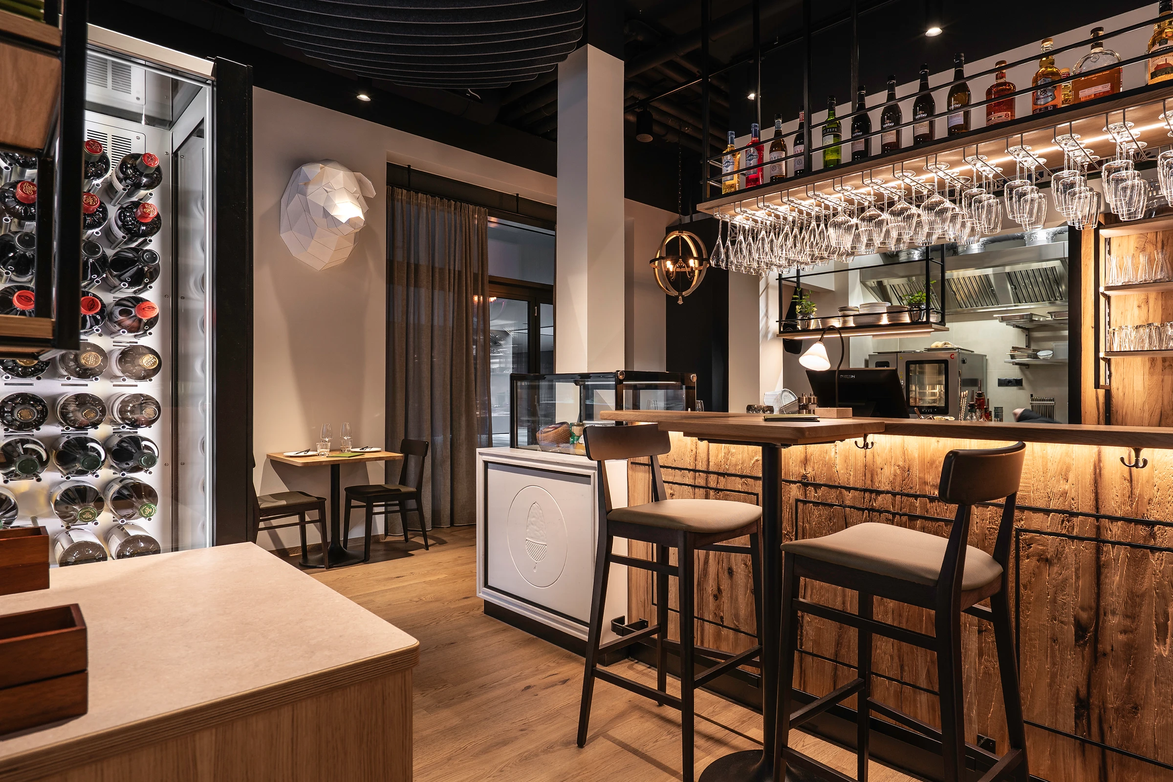 Bar des Paco Ribera mit beleuchtetem Weinkühlschrank, Barhockern am Naturholztresen, hängenden Weingläsern, offener Küche, weißer Skulptur an der Wand und dunkler Akustikdecke.