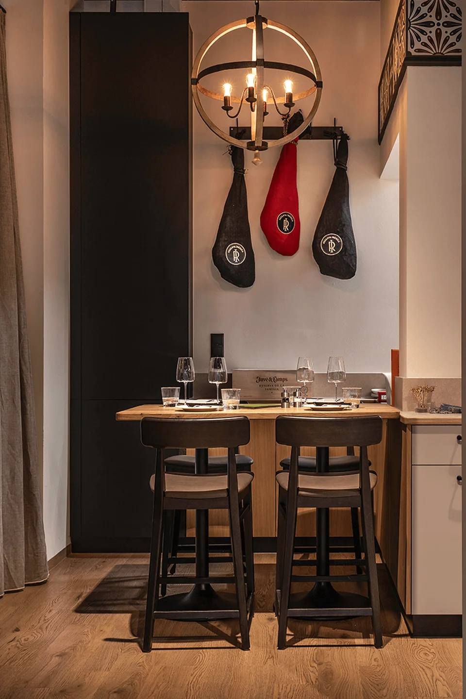 Sitzbereich des Paco Ribera mit zwei hängenden Schinken mit Logo-Aufdruck, goldener Kugelleuchte, gedecktem Holztisch, schwarzen Barhockern und grauem Vorhang als Raumtrenner.