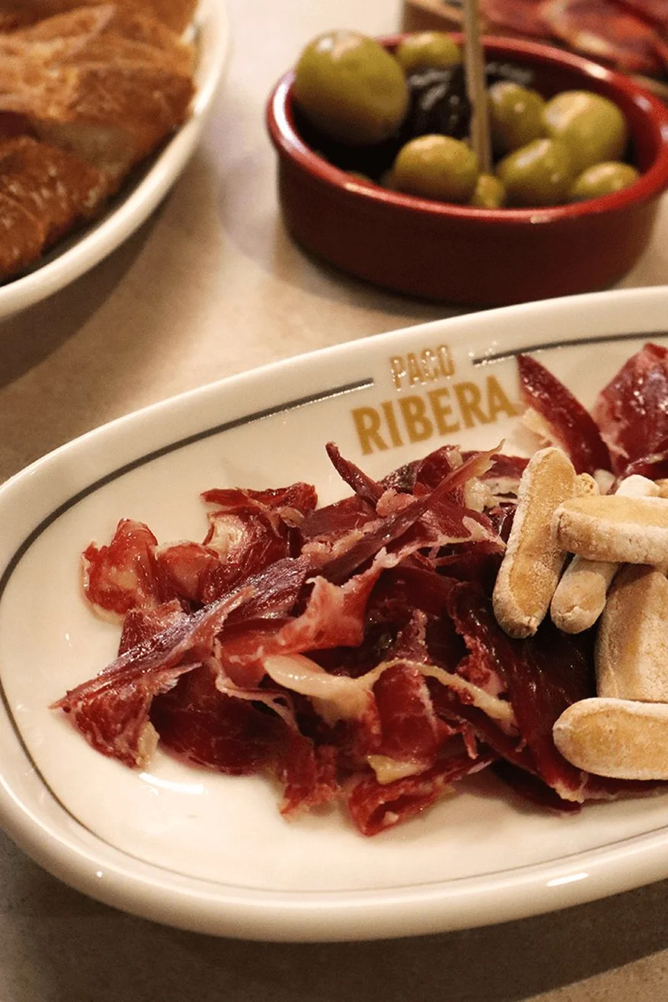 Tapas-Auswahl im Paco Ribera auf dem markanten Logo-Teller mit hauchdünn geschnittenem Jamón, Grissini, Oliven in Tonschale und Weißbrot im Hintergrund.