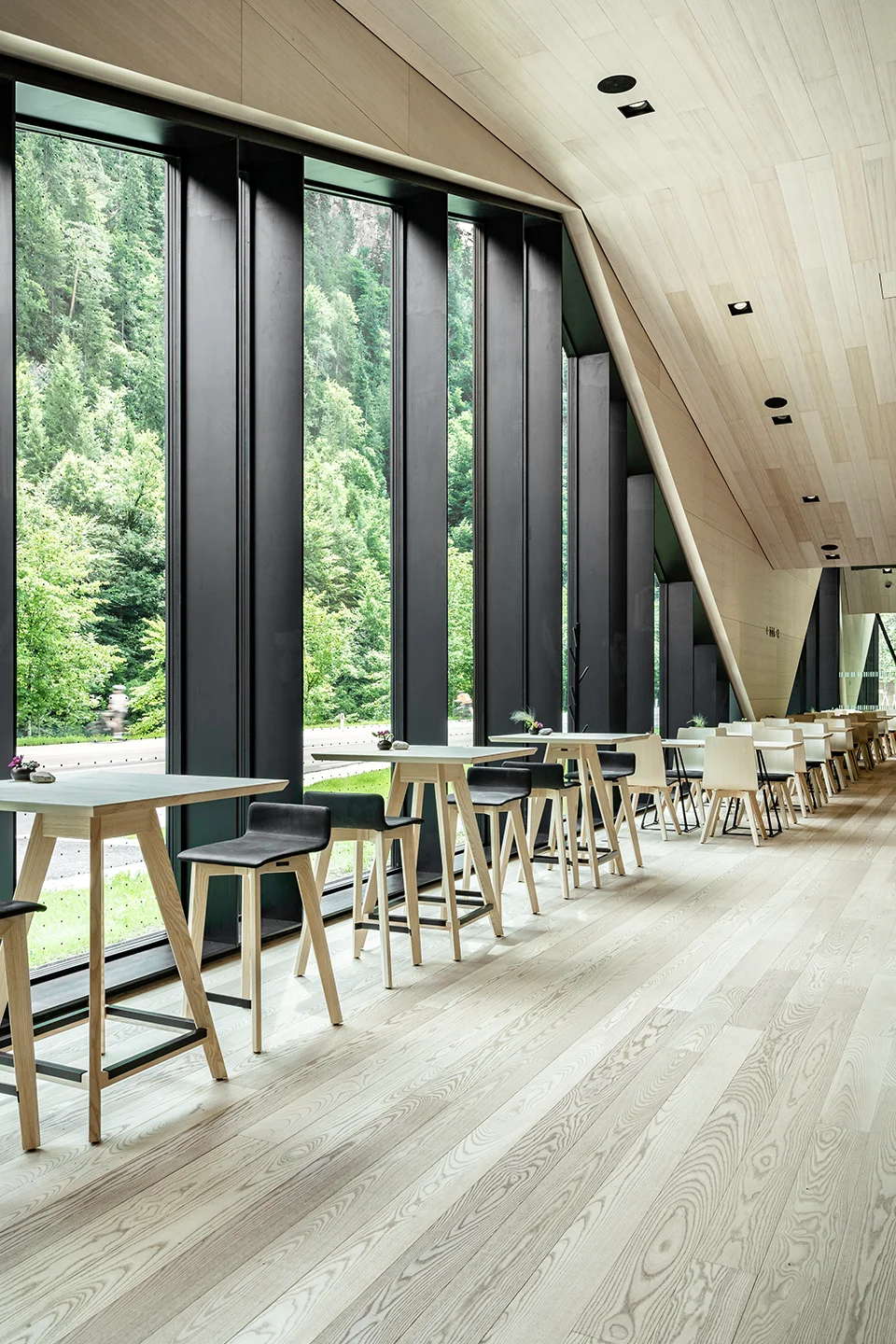 Hochbereich des Fort Kniepass Cafés in Unken mit langen Stehtischen, schwarzen Barhockern aus Eschenholz, raumhoher Glasfront mit Waldblick und schräger Holzdecke