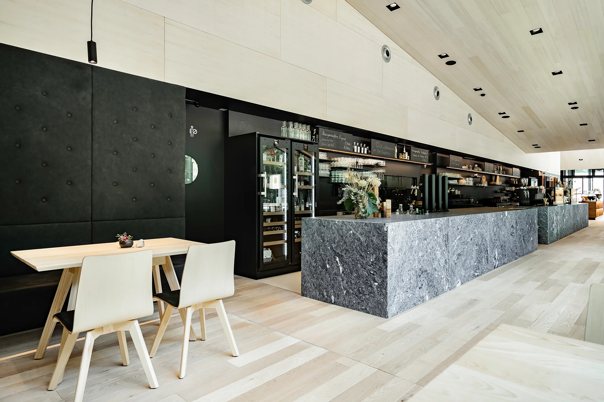 Innenansicht des Fort Kniepass Cafés in Unken mit Granittresen, Weinkühlschrank, schwarzer Bareinrichtung, Holzstühlen und schräger Holzdecke