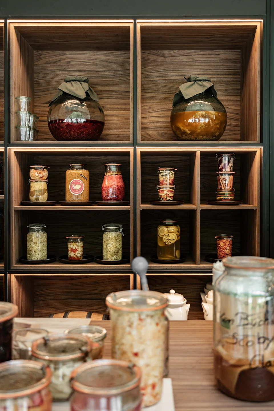 Detail des beleuchteten Fermentationsregals im Fort Kniepass mit großen Kugelgläsern, verschiedenen Einmachgläsern mit fermentierten Produkten in Nussbaumholzfächern