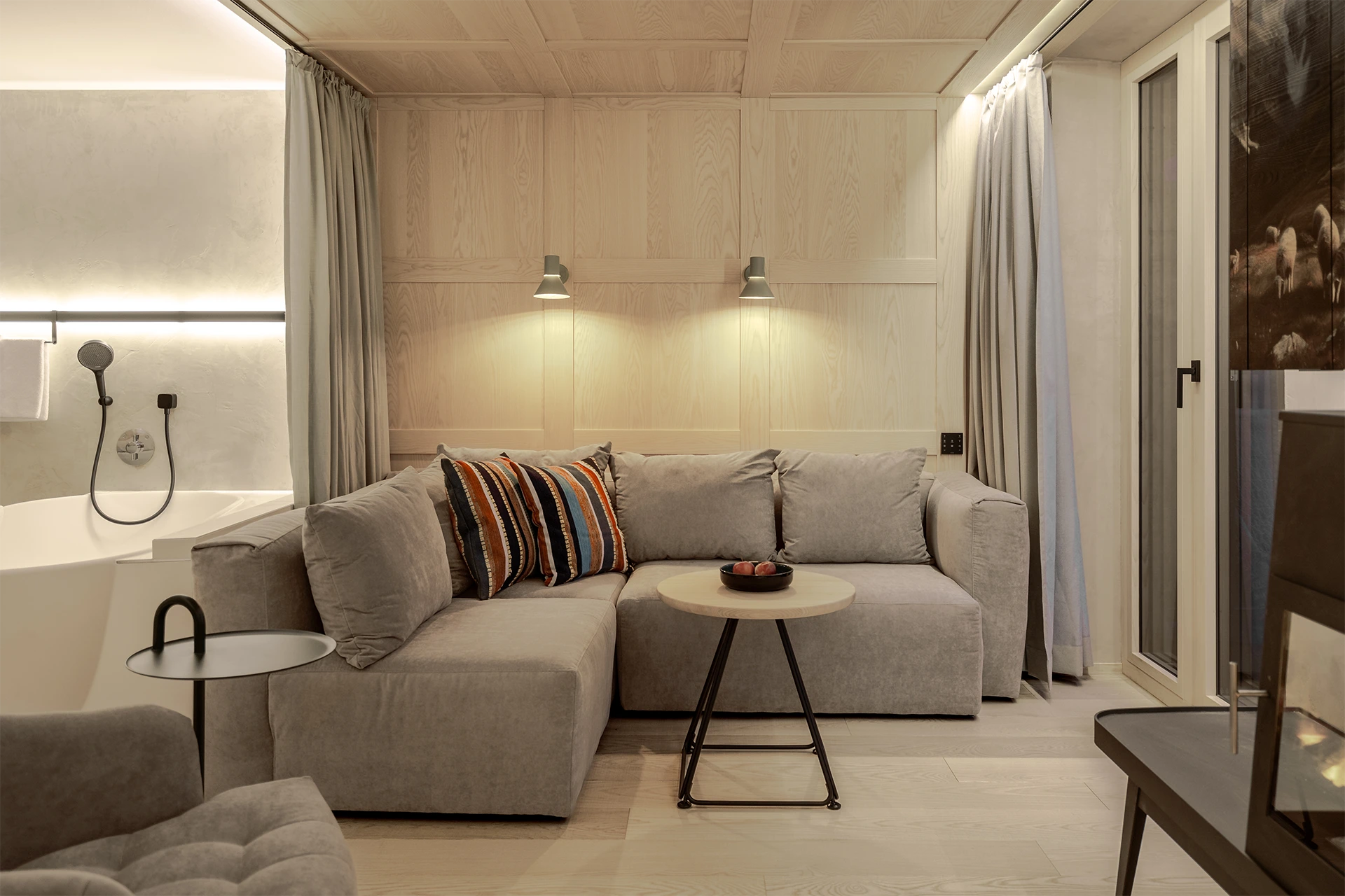 Wohnbereich mit grauem Ecksofa, bunten Akzentkissen, Holztisch und einer freistehenden Badewanne im Hintergrund.