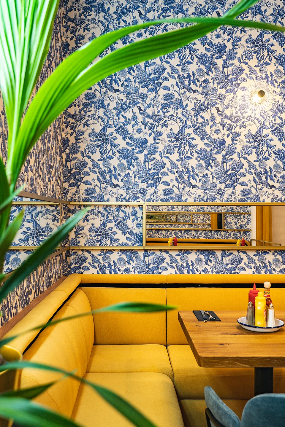 Gemütliche Sitz-Ecke mit einer senfgelben Eck-Polsterbank vor einer auffälligen blauen Blumentapete und einem länglichen Wandspiegel.