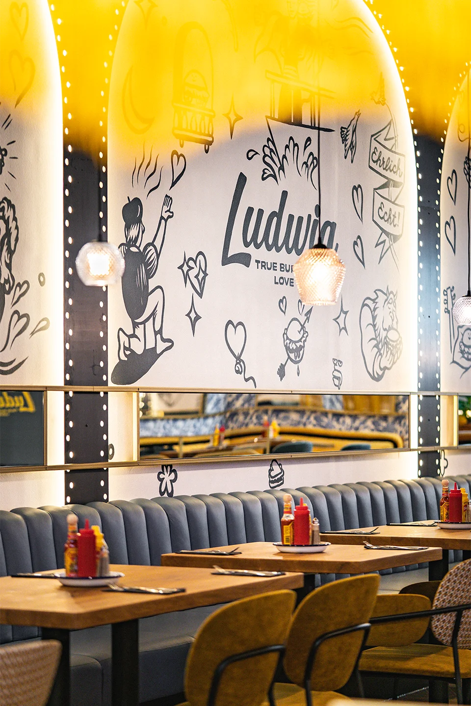 Sitzbereich mit blauen Diner-Bänken aus Leder und Holztischen vor beleuchteten Rundbögen mit handgezeichneten Illustrationen zum Thema Burger.