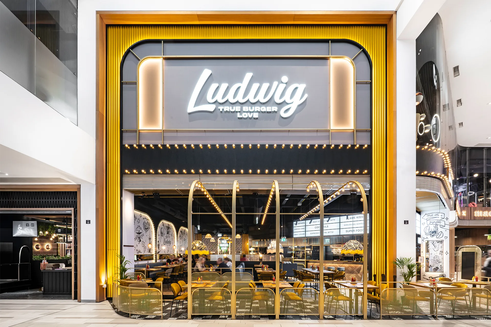 Frontale Eingangsfassade des Restaurants Ludwig mit großem Leuchtlogo, goldfärbigen seitlichen Wellblech und einem auffälligen schwarz-weiß gemusterten Fliesenboden.