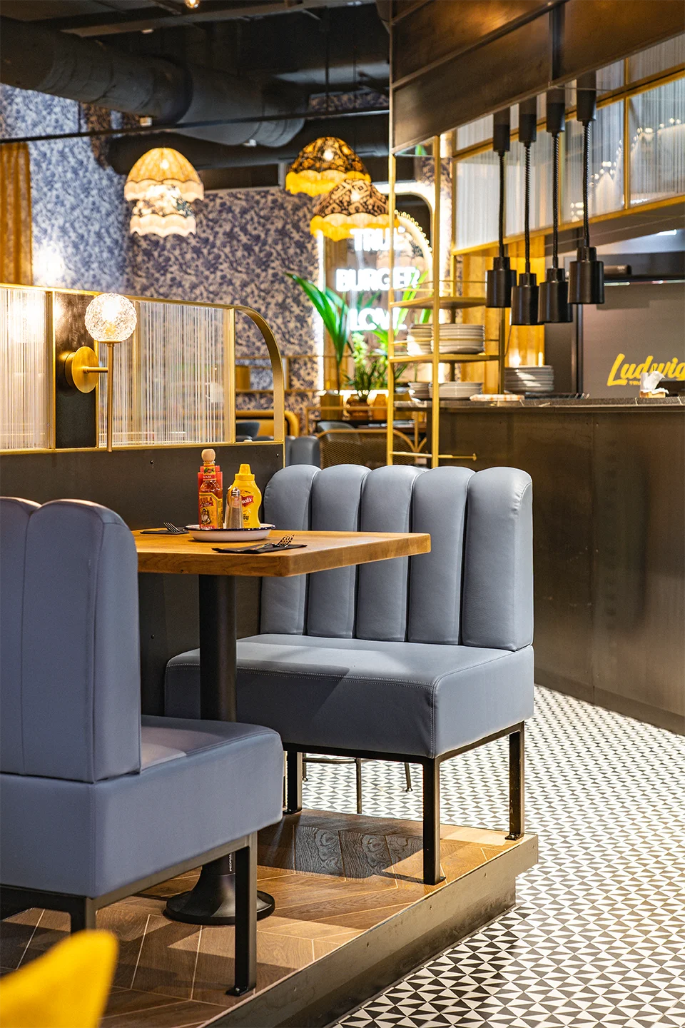 Detailansicht einer gemütlichen Diner-Sitzbank mit hellblauen Leder-Tapezierung an einem Holztisch, getrennt durch eine elegante Raumtrennung aus Riffelglas und Messing.