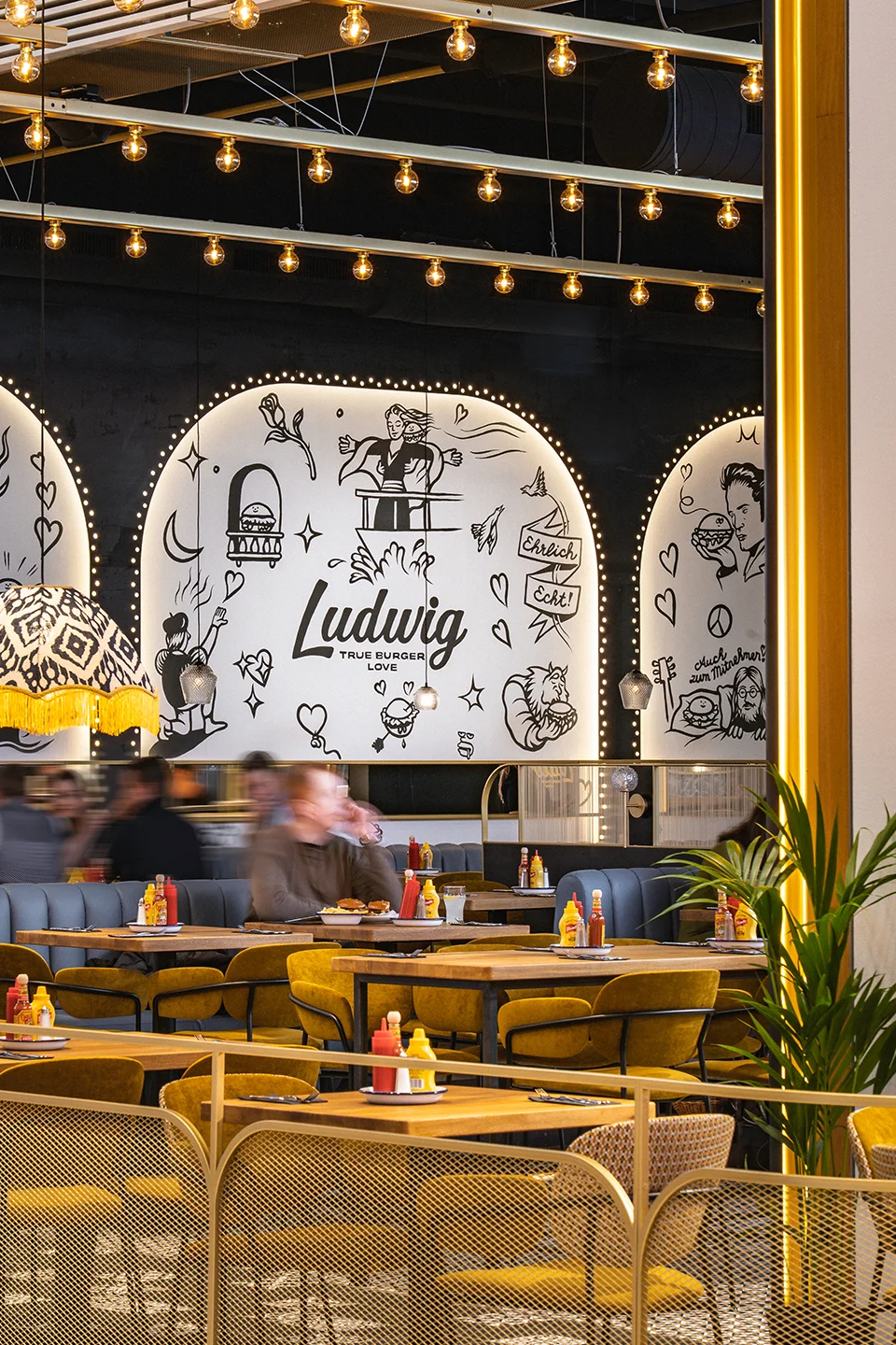 Belebter Sitzbereich im Restaurant mit senfgelben Polsterstühlen, Glühbirnenband an der Decke und einer großen, kreativen Wandbemalung im Hintergrund.