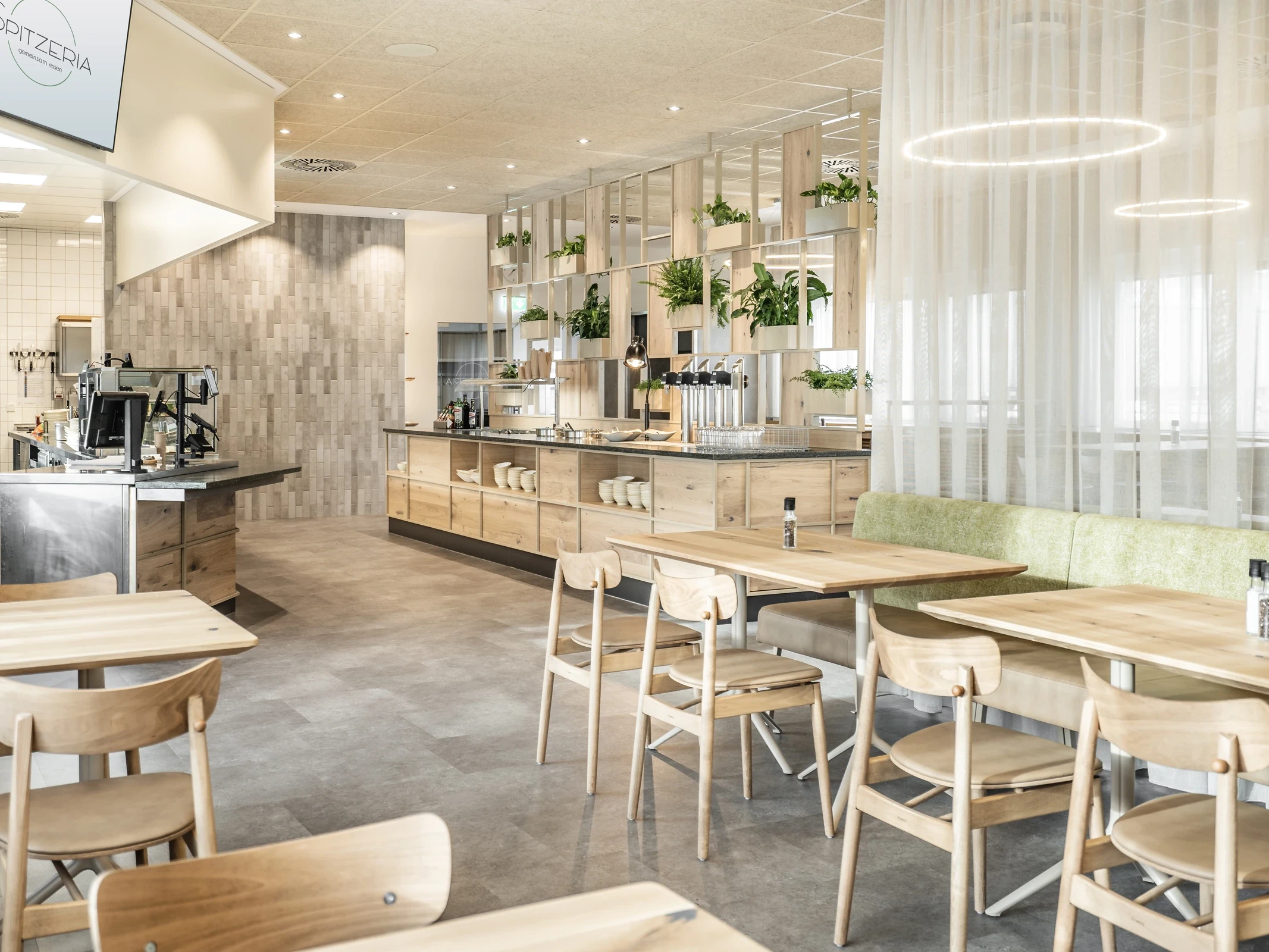 Betriebsrestaurant Spitzeria in Attnang mit Warmhaltestrahler, Tellern auf der Theke und bepflanztem Holzregal im Hintergrund.
